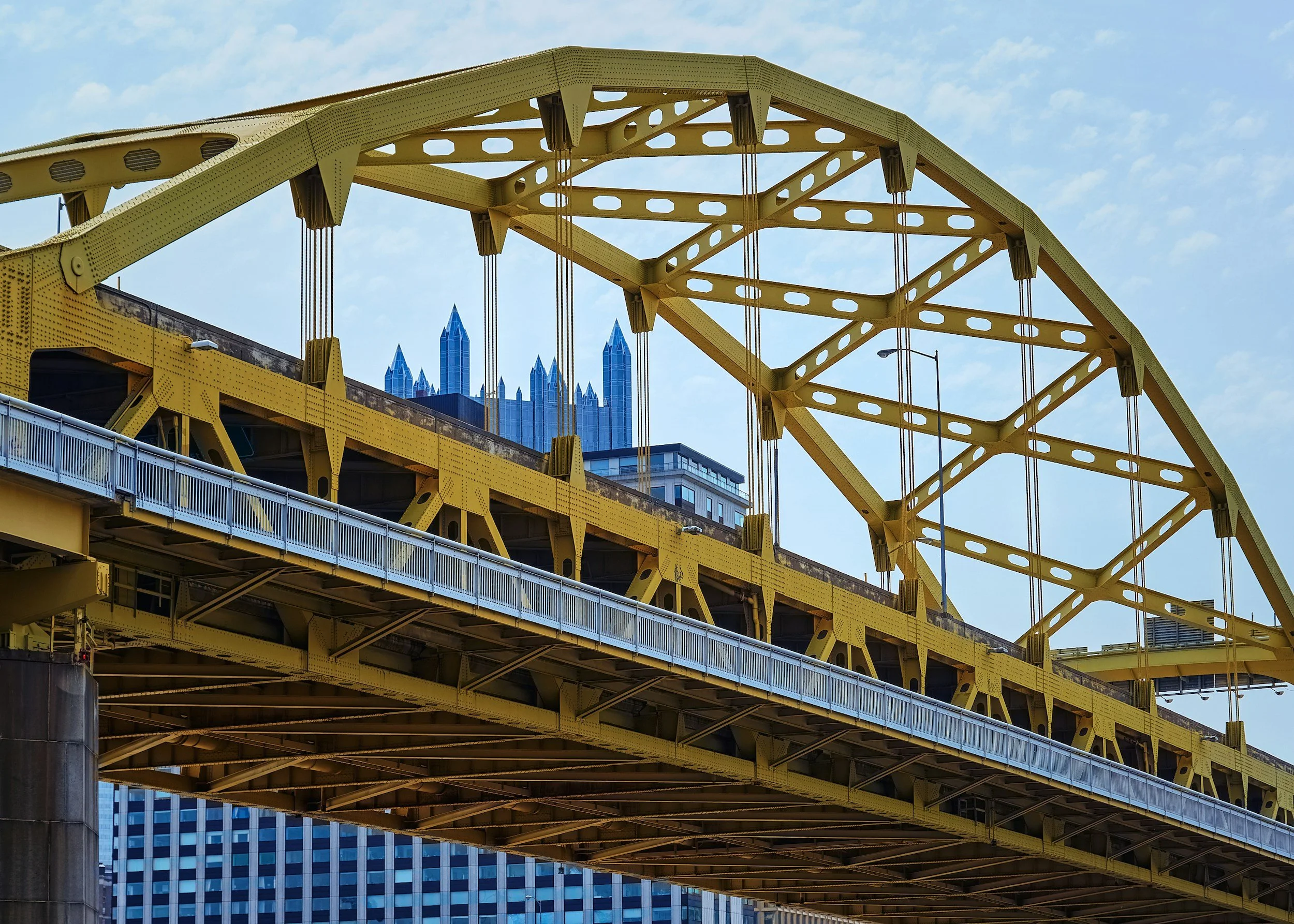A yellow steel bridge with a city skyline in the background, including tall buildings with pointed tops.