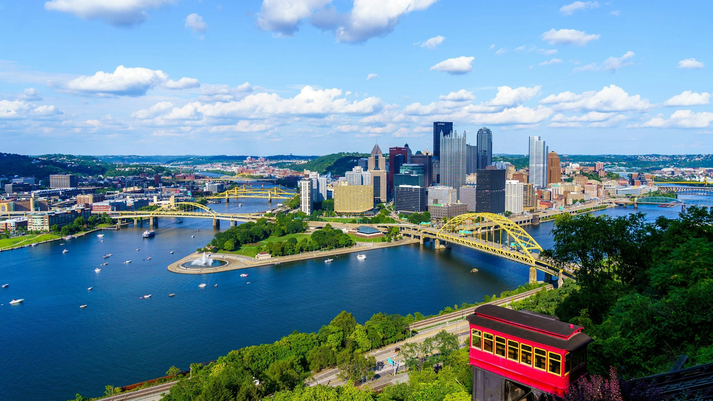 A panoramic view of Pittsburgh, Pennsylvania, showcasing the skyline with tall buildings, yellow bridges crossing the river, and boats on the water, with a red funicular railway in the foreground and lush greenery around.