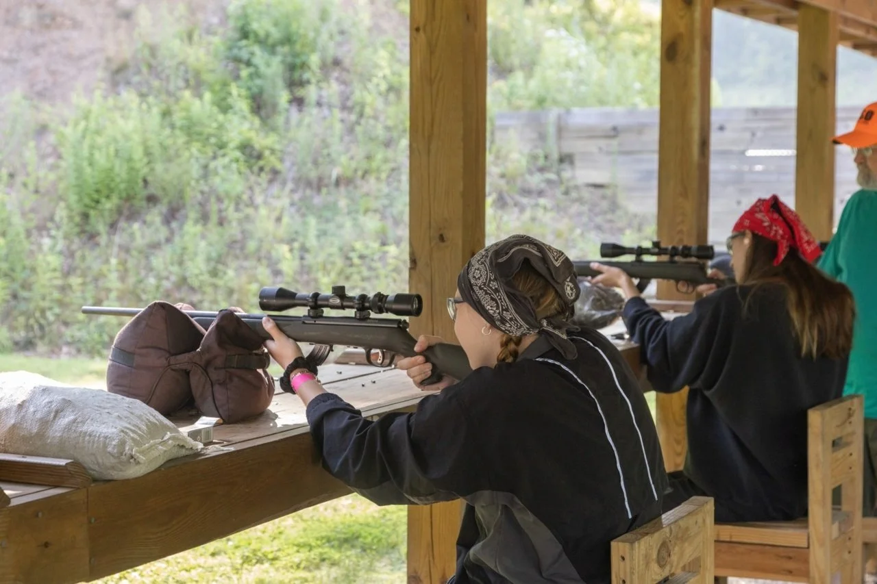Three people at a shooting range, aiming rifles with scopes under a wooden shelter. Two women with bandanas on their heads and an older man with a beard and orange cap are practicing shooting.