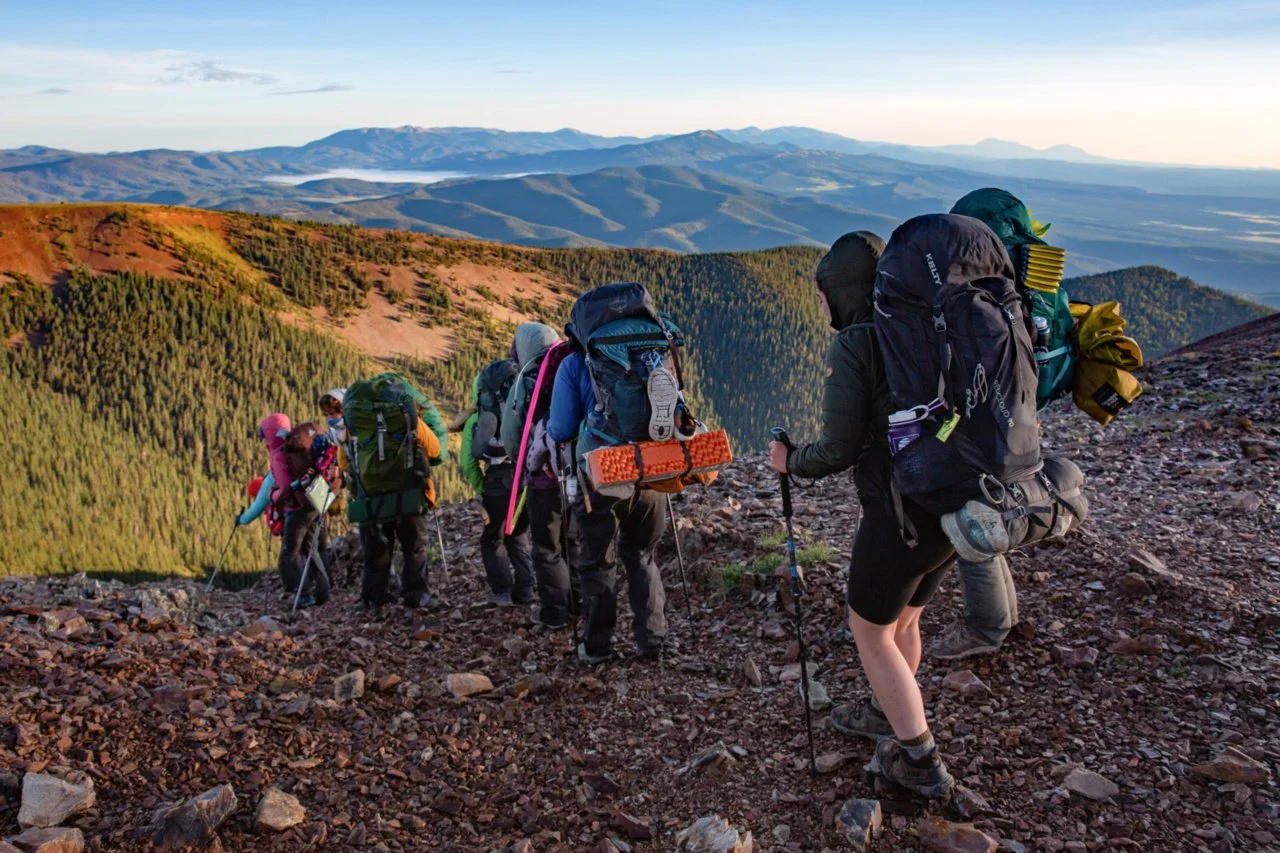Group of hikers trekking up a mountain trail with scenic mountain ranges in the background during sunrise or sunset.