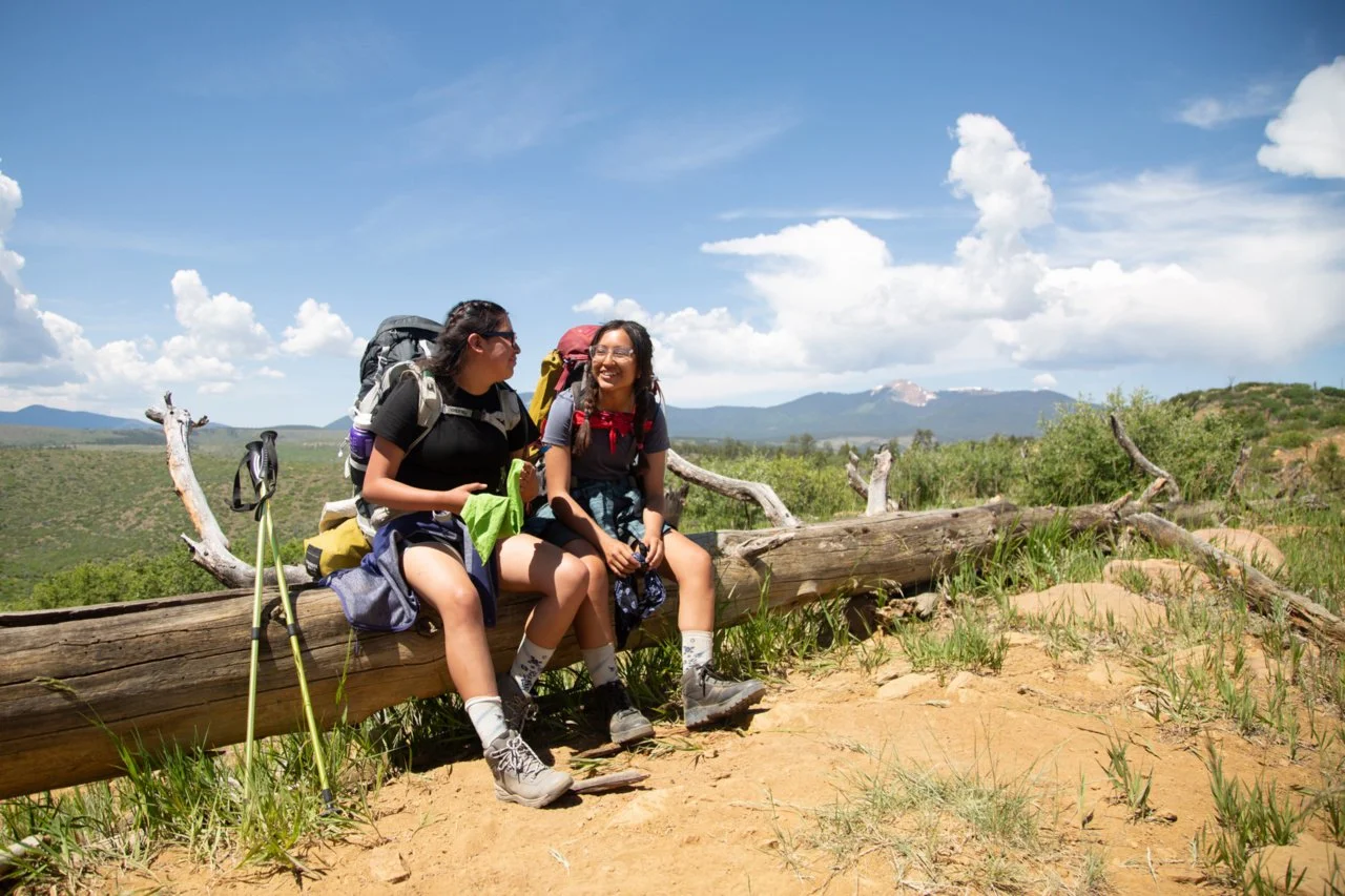 Two young women with backpacks sitting on a fallen log outdoors, smiling and talking amidst green grass and open skies with clouds, mountains in the distance.