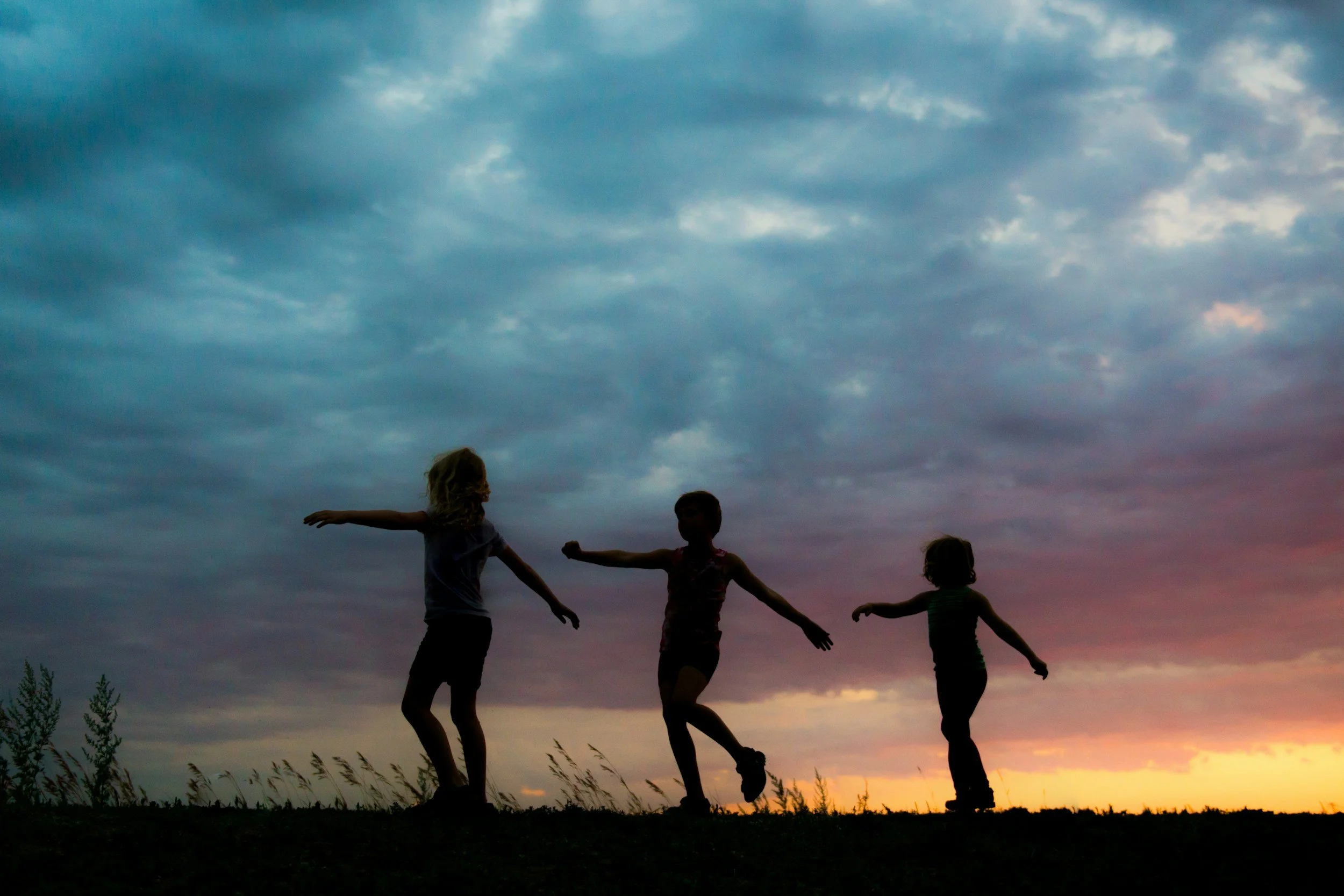 Silhouettes of three children playing outdoors during sunset with a cloudy sky.