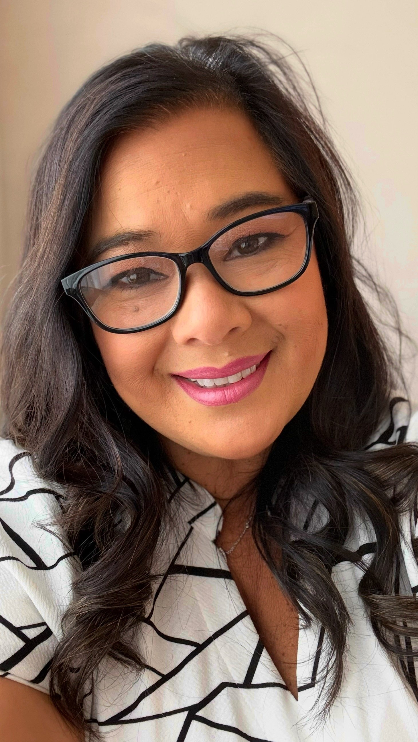 Close-up of a woman with long wavy dark hair, black glasses, pink lipstick, and a white blouse with black abstract lines, smiling.