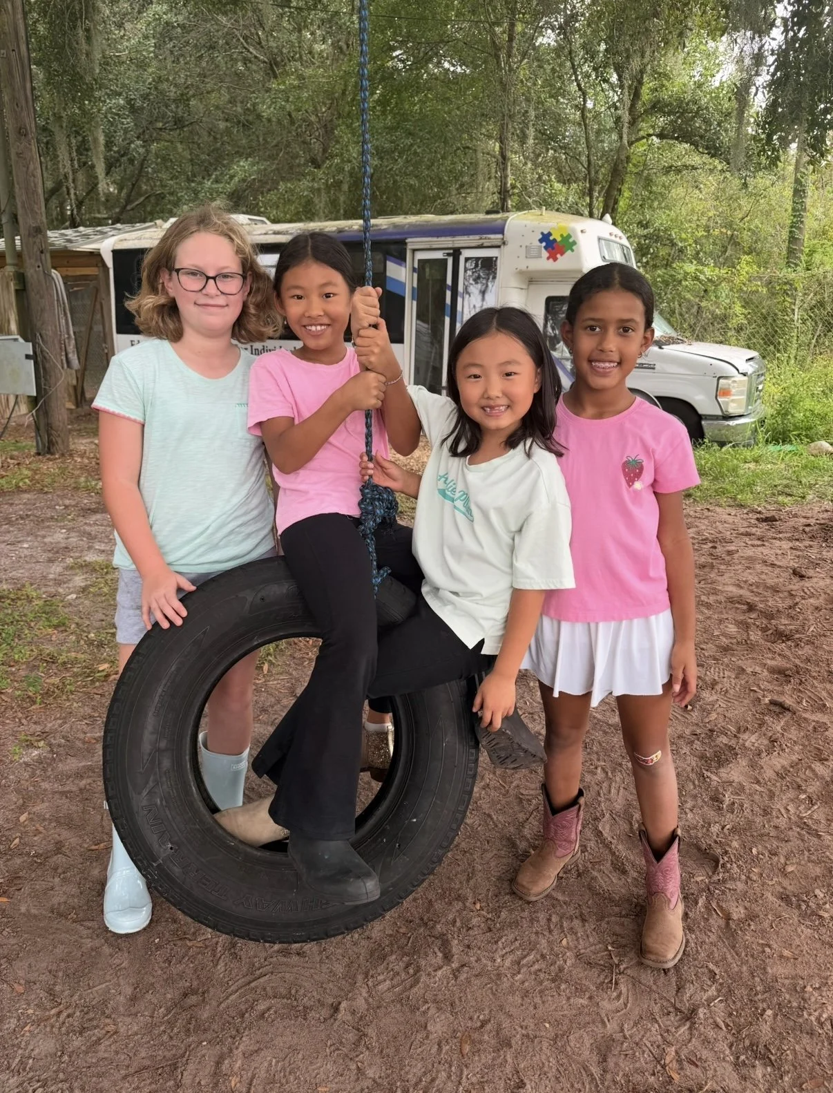 Four young girls smiling and standing together outdoors around a tire swing, with a forest background and a white van in the distance.