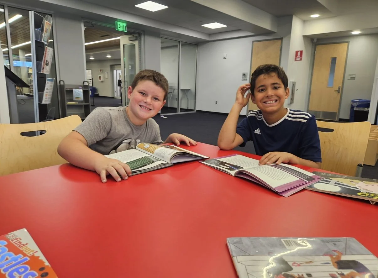 Two boys sitting at a red table in a library or study area, reading and smiling, with books open in front of them.