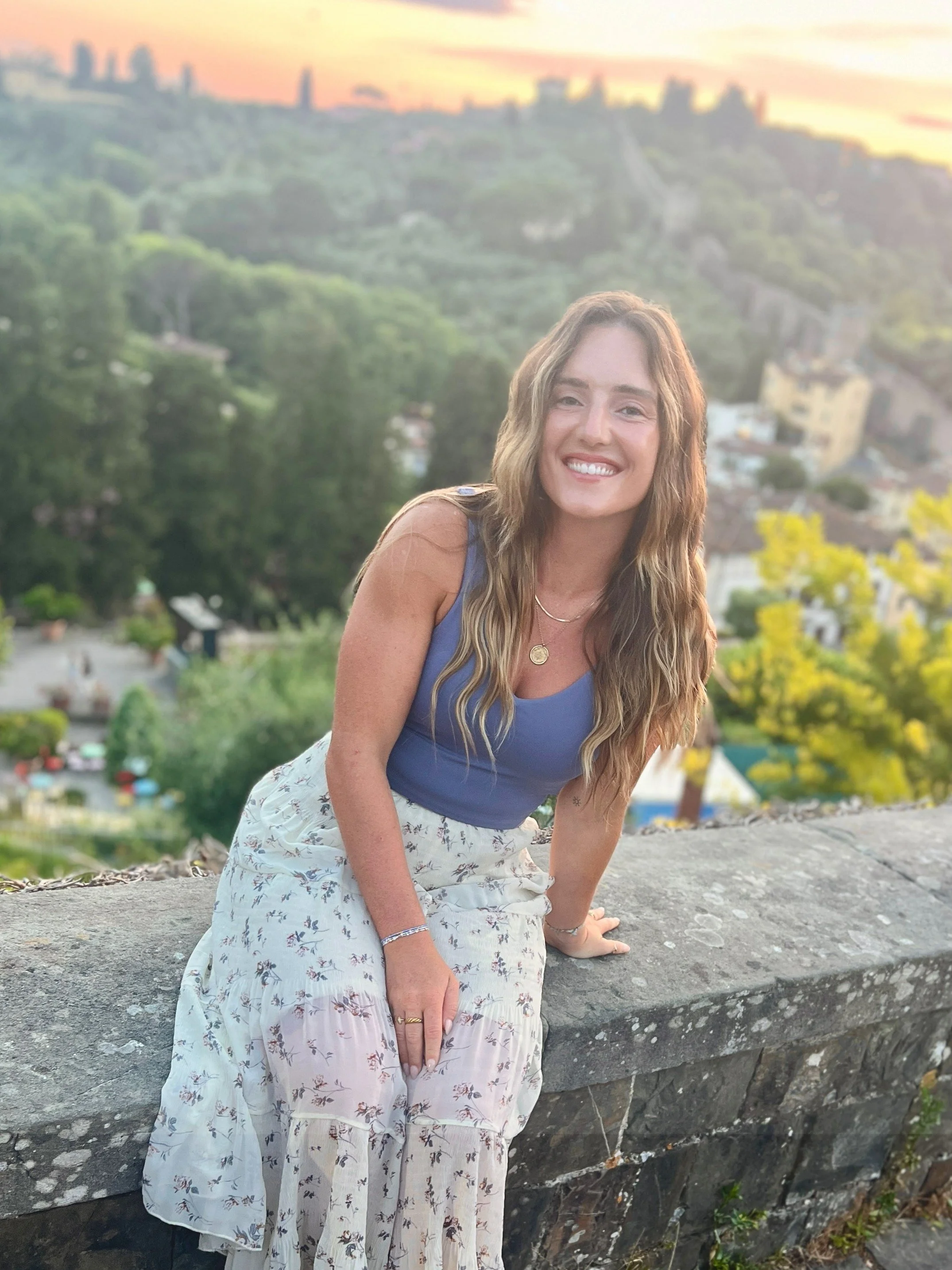 Smiling woman with long wavy hair sitting on a stone wall with a city and green hills in the background during sunset.