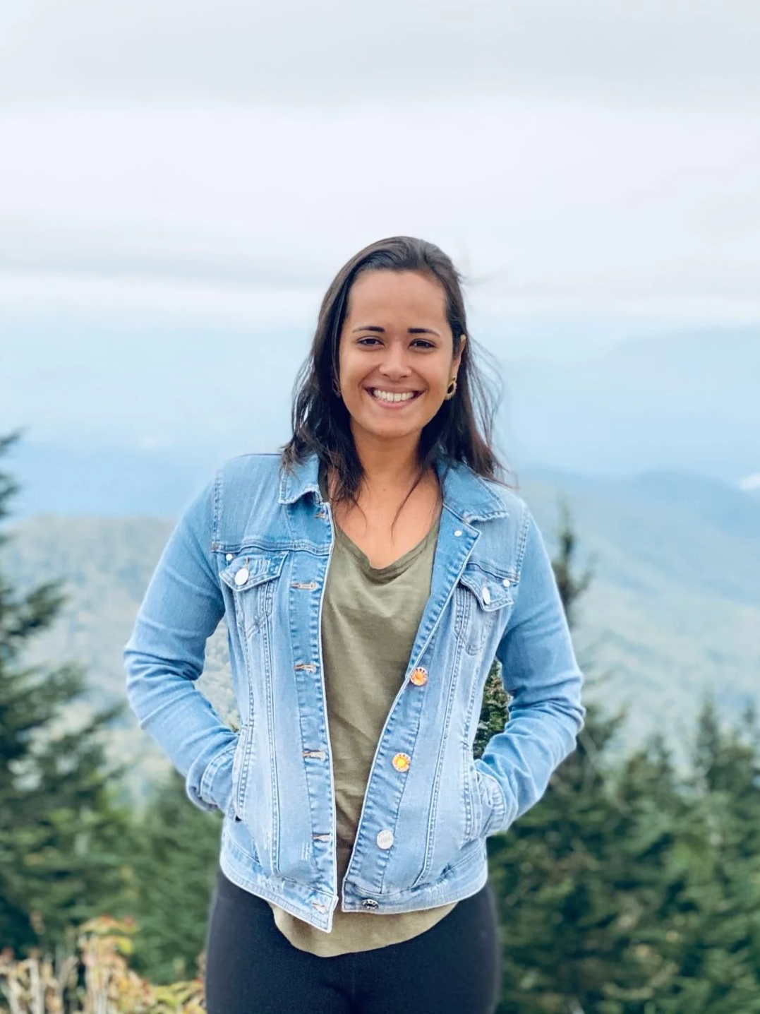 A young woman smiles outdoors in front of a mountain landscape, wearing a denim jacket and black pants.