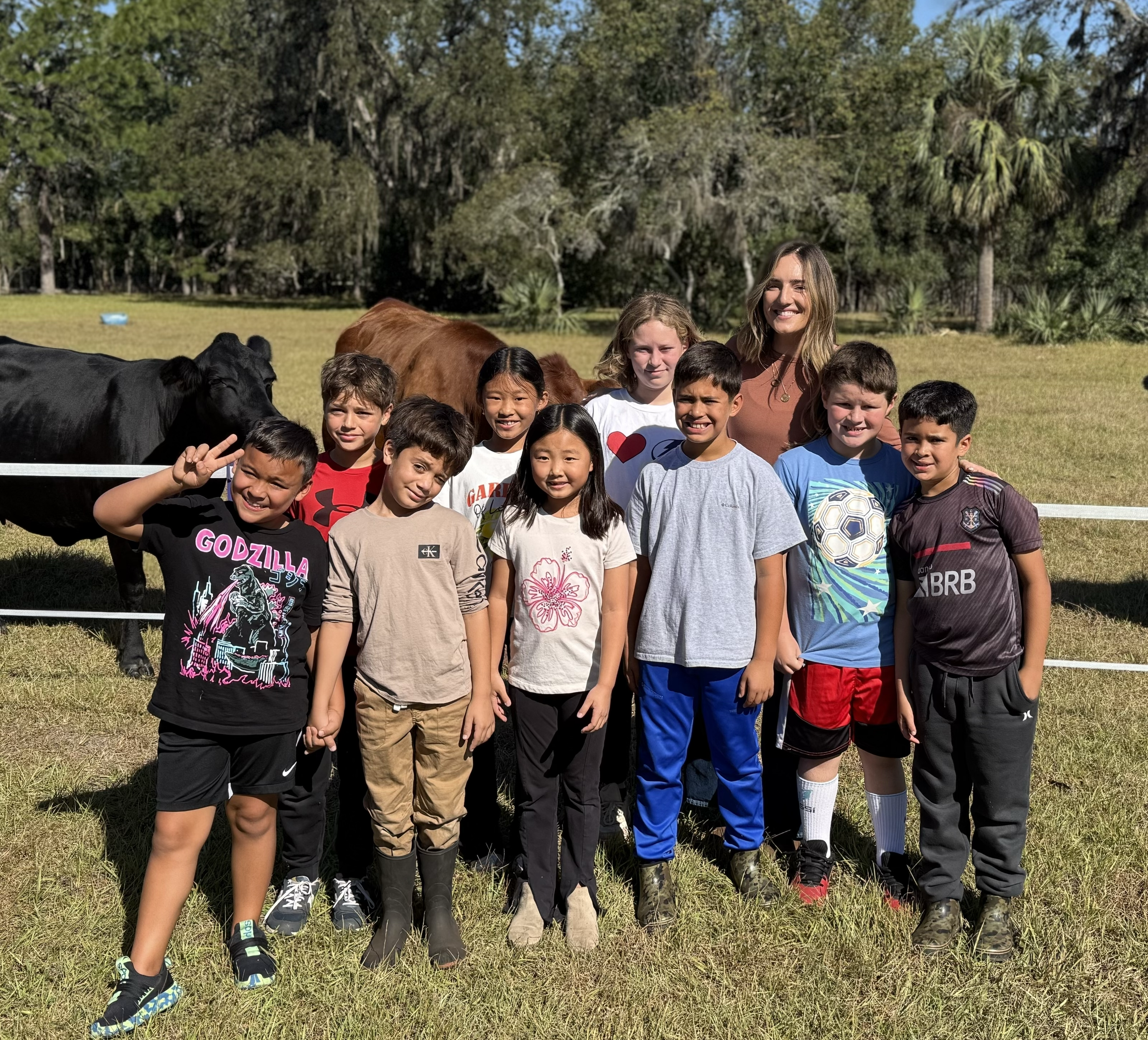 Group of children and a woman standing outdoors on grass, with cows and trees in the background.