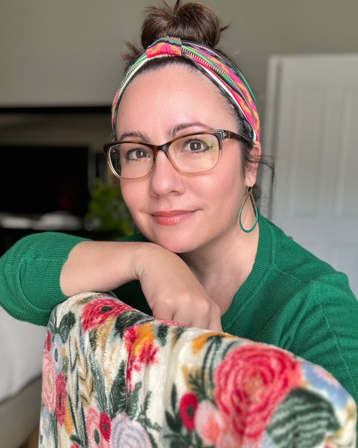 A woman with glasses and a colorful headband, resting her chin on her hand, smiling gently. She is wearing a green top and a floral sleeve with pink, red, orange, and purple flowers.
