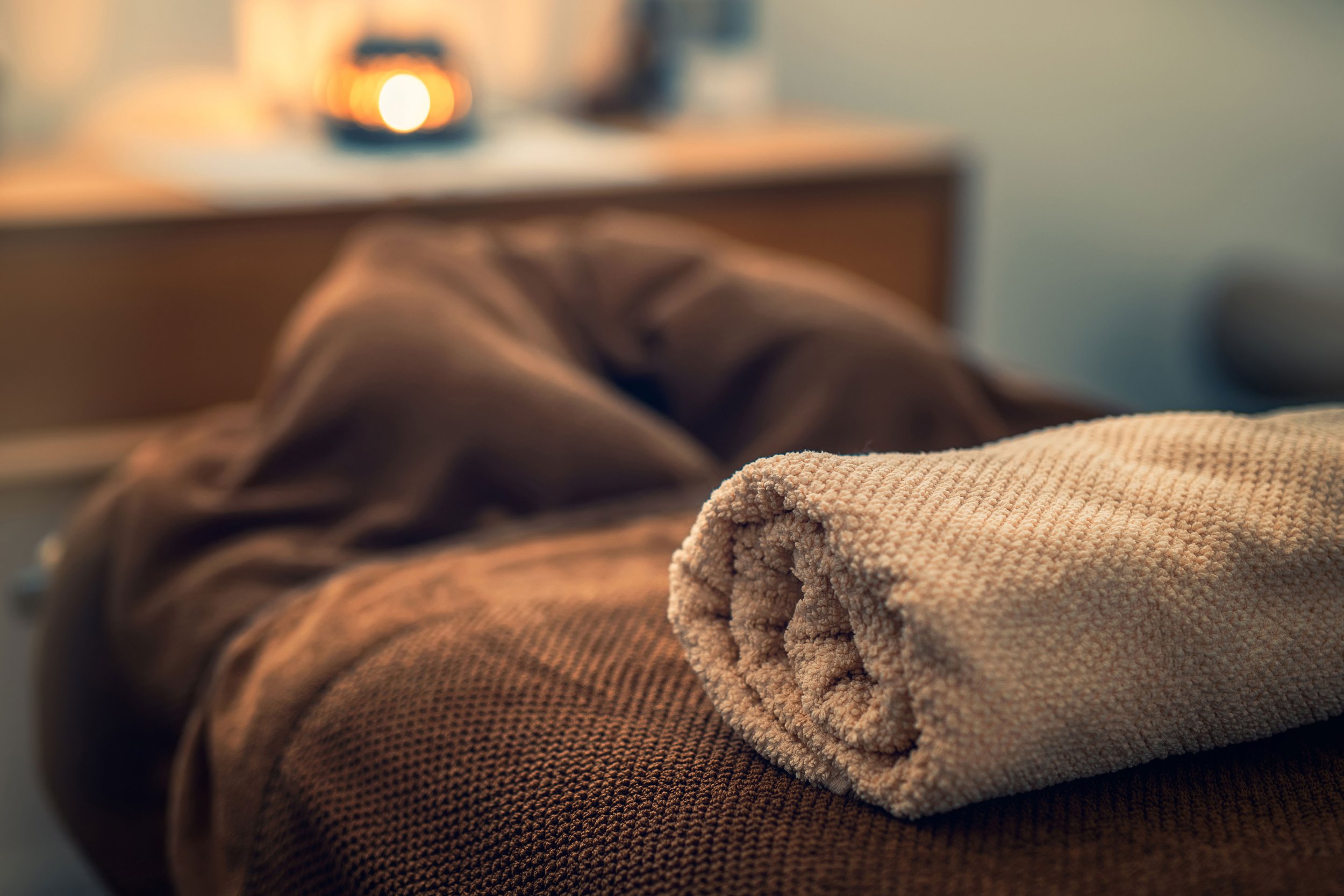 Rolled beige towel on a bed with brown bedding, in a softly lit room.