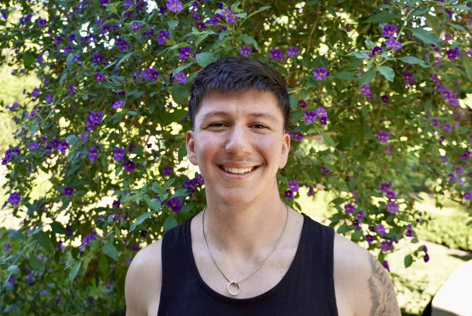 A smiling young man with short dark hair, wearing a black tank top and a circular pendant necklace, stands outdoors in front of a large bush with purple flowers and green leaves.
