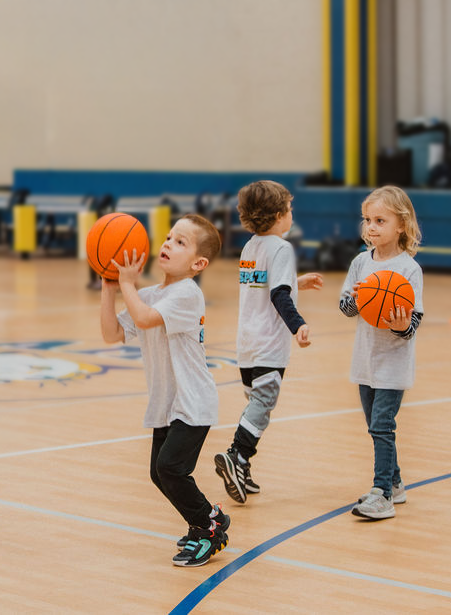 Three young children playing with basketballs on a gymnasium basketball court.