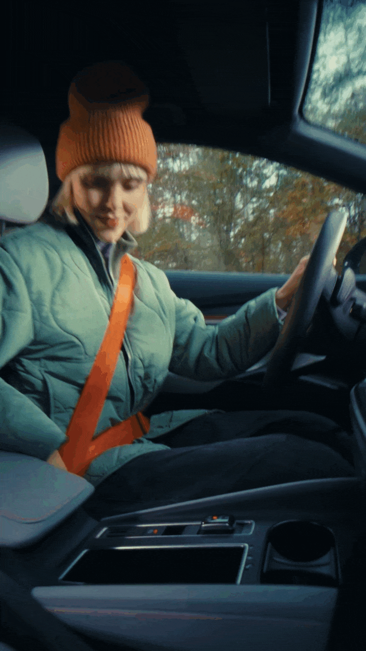 ŠKODA ELROQ. A woman in a green jacket and orange beanie sitting in the driver's seat of a car, smiling and looking at the camera, with trees visible outside the window. Driving on a forest road.