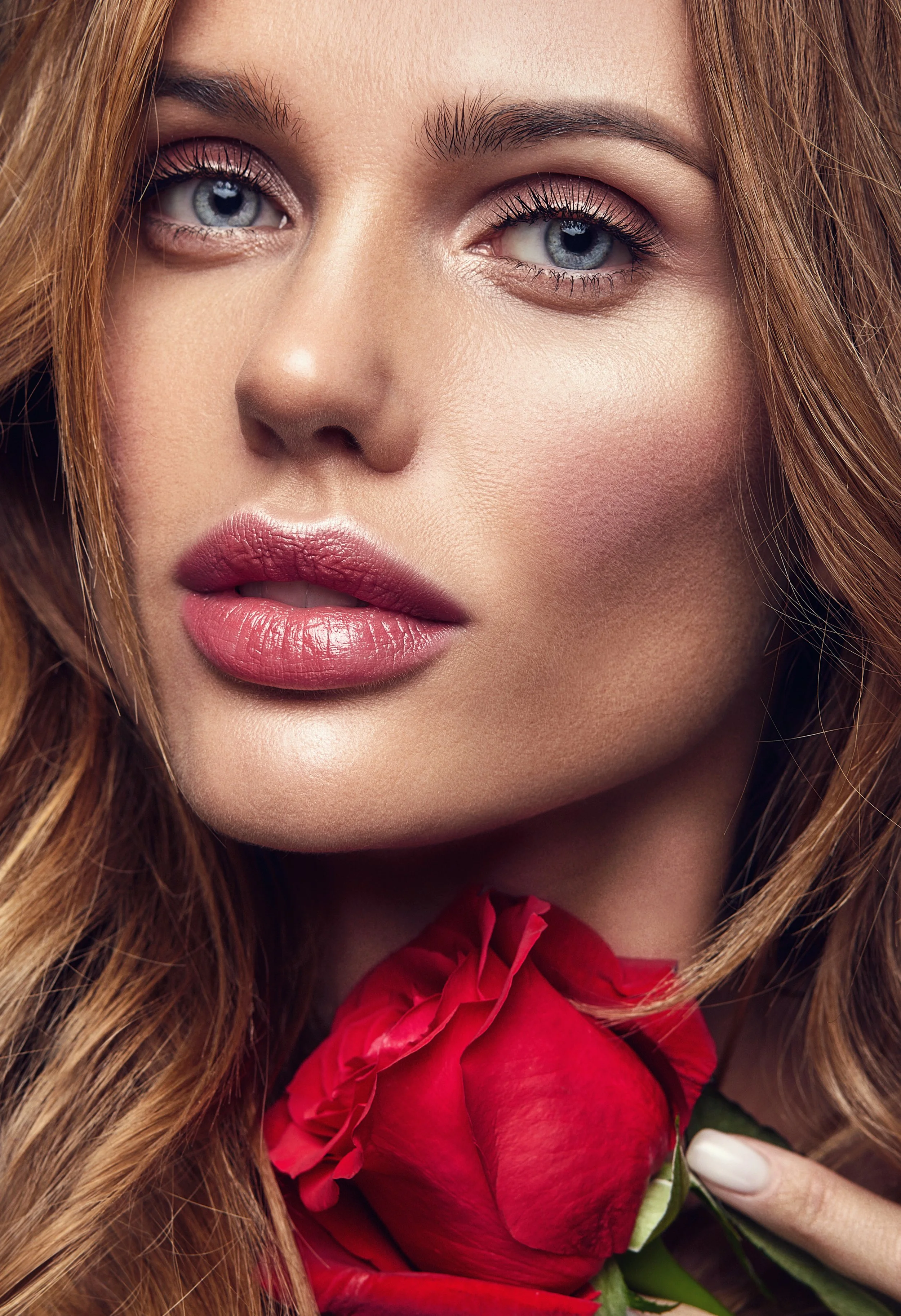 A close-up of a woman with fair skin, blue eyes, lifted eyebrows, red hair, pink lipstick, and eye makeup, holding a red rose near her chin.