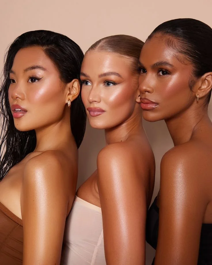 Three women with different ethnicities and skin tones posing closely together. They have styled hair and are wearing strapless clothing, with earrings, against a plain beige background.