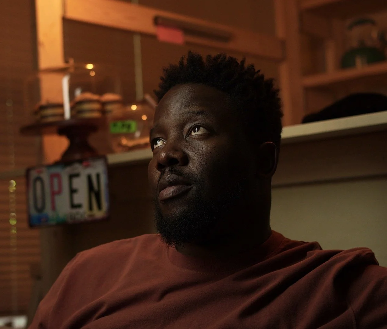 A man with dark skin, a beard, and short curly hair looking upward, sitting in a dimly lit cafe with an 'OPEN' sign in the background.