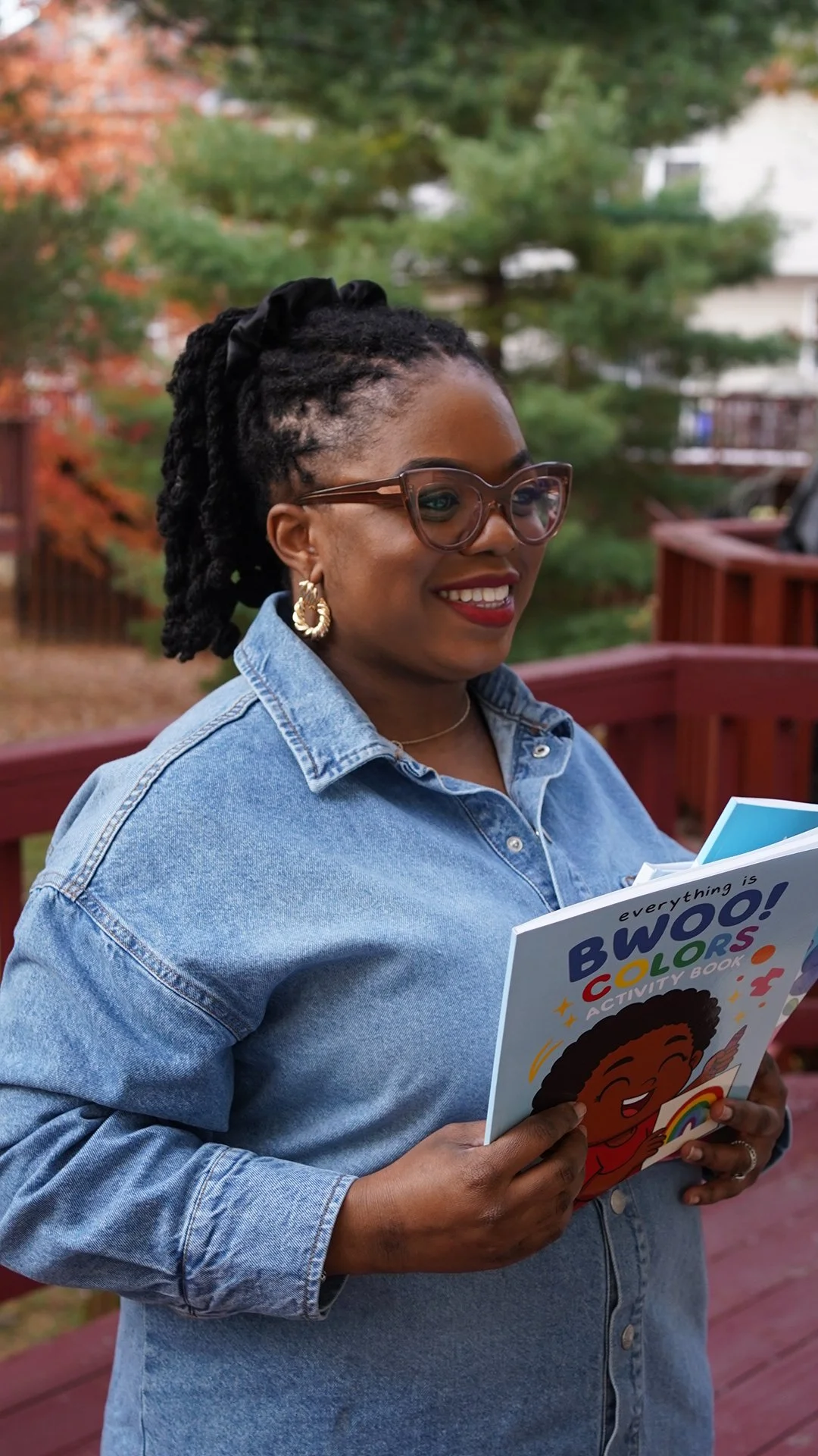 Woman with glasses and earrings reading a colorful children's activity book outdoors on a wooden deck, with trees in the background.