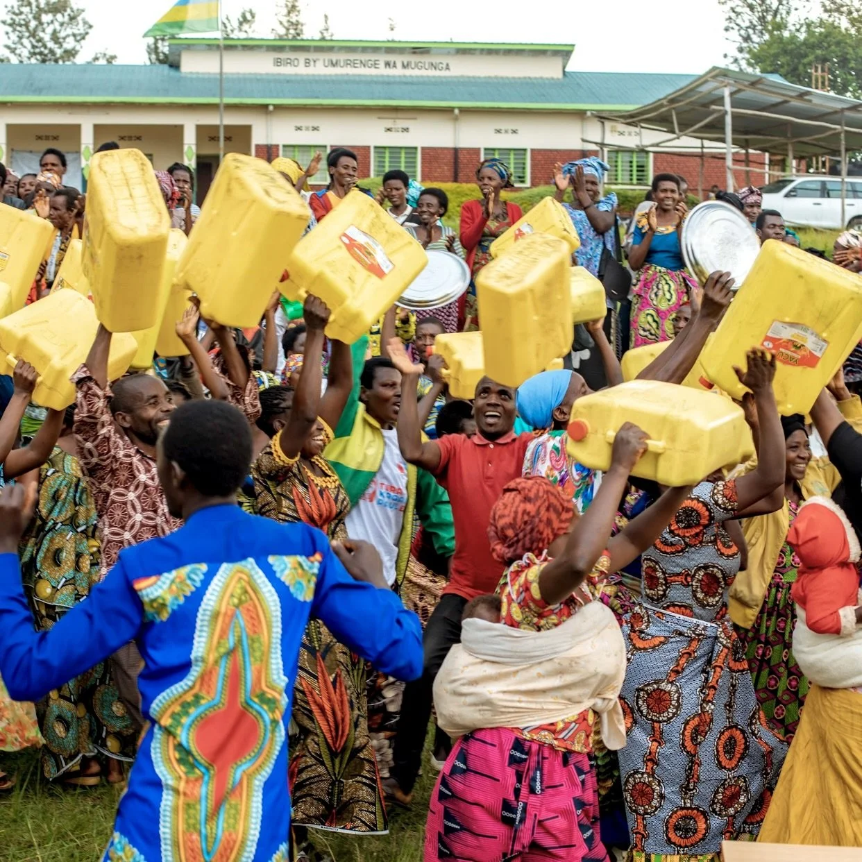 Crowd of people celebrating outdoors, many holding yellow water jerry cans and metal trays, with a school building in the background.
