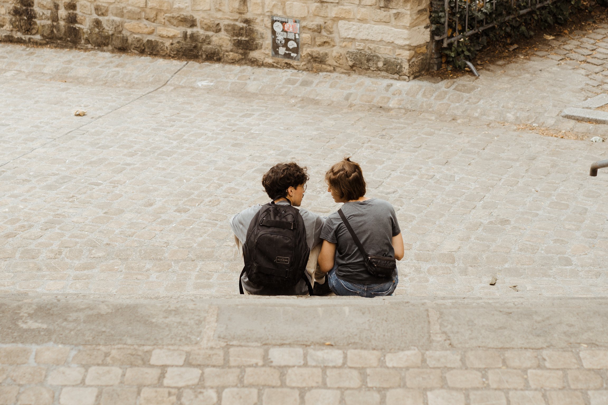 Two people sitting on the cobblestone ground, facing each other, with backpacks, in front of a brick wall.