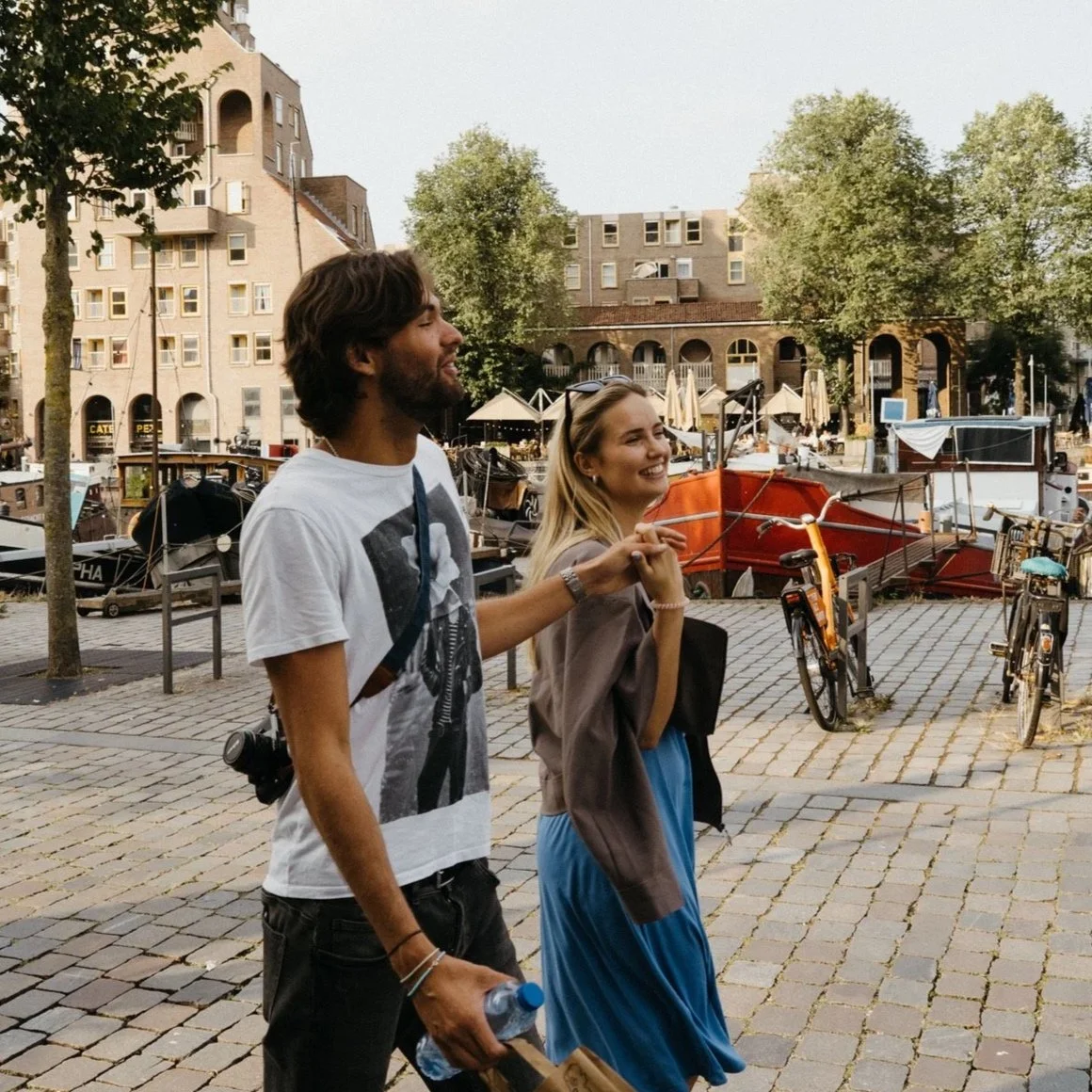 A man and woman walking along a cobblestone pathway near a marina with boats docked, trees, and multi-story buildings in the background, smiling and enjoying a casual outdoor moment.