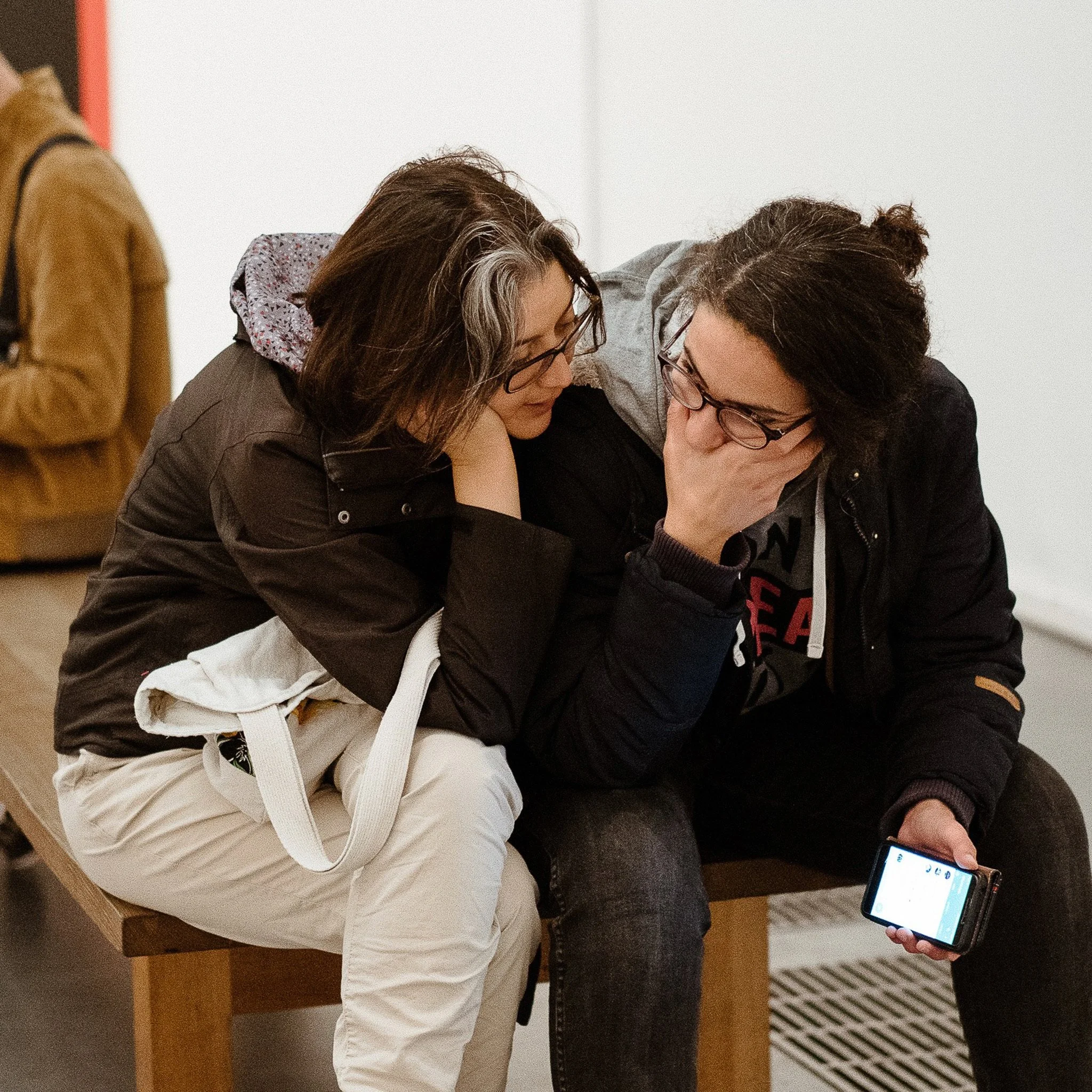Two women with glasses leaning close together, looking at something on a cellphone, with a third person partially visible on the left in a brown jacket.