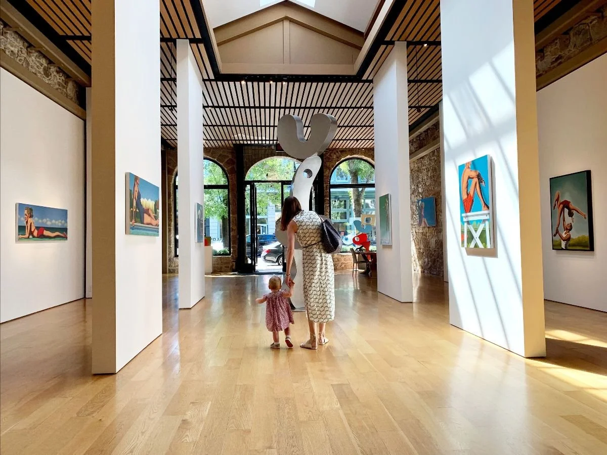 Woman and small girl walking in an art gallery with paintings on white walls, large sculpture hanging from the ceiling, and large windows showing street view outside.