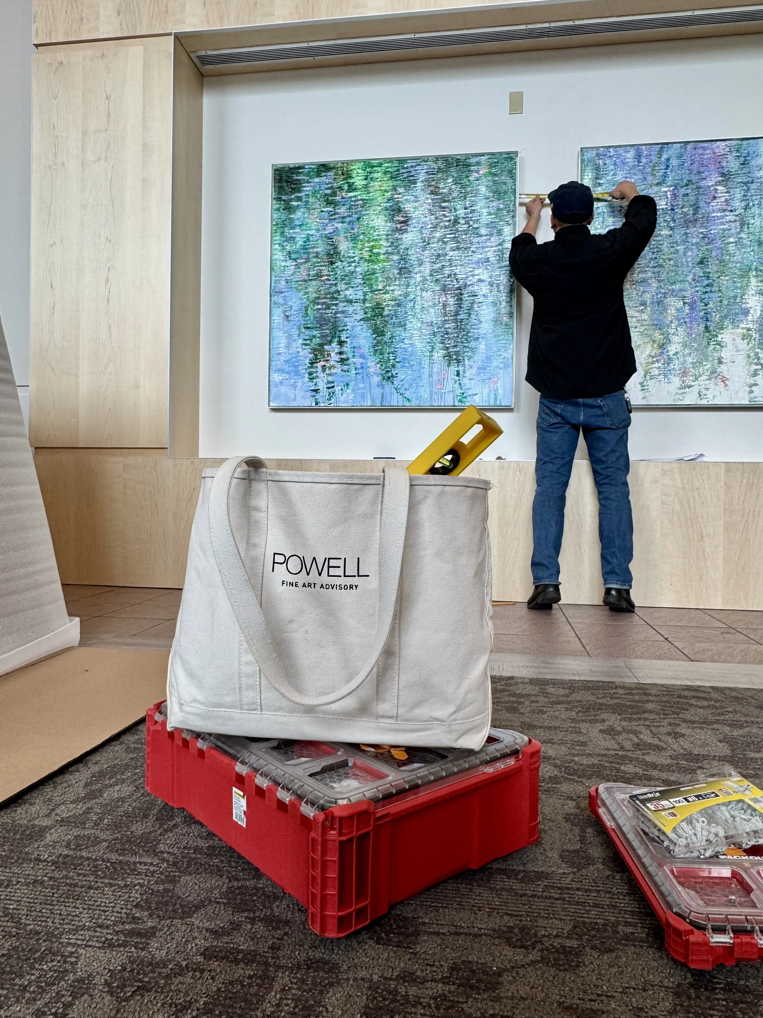 A person hanging or installing artwork on a gallery or museum wall. In the foreground, there is a white tote bag with the text 'Powell Fine Art Advisory' on it, placed on a red plastic crate. Next to the tote bag, there is a yellow level and a plasti