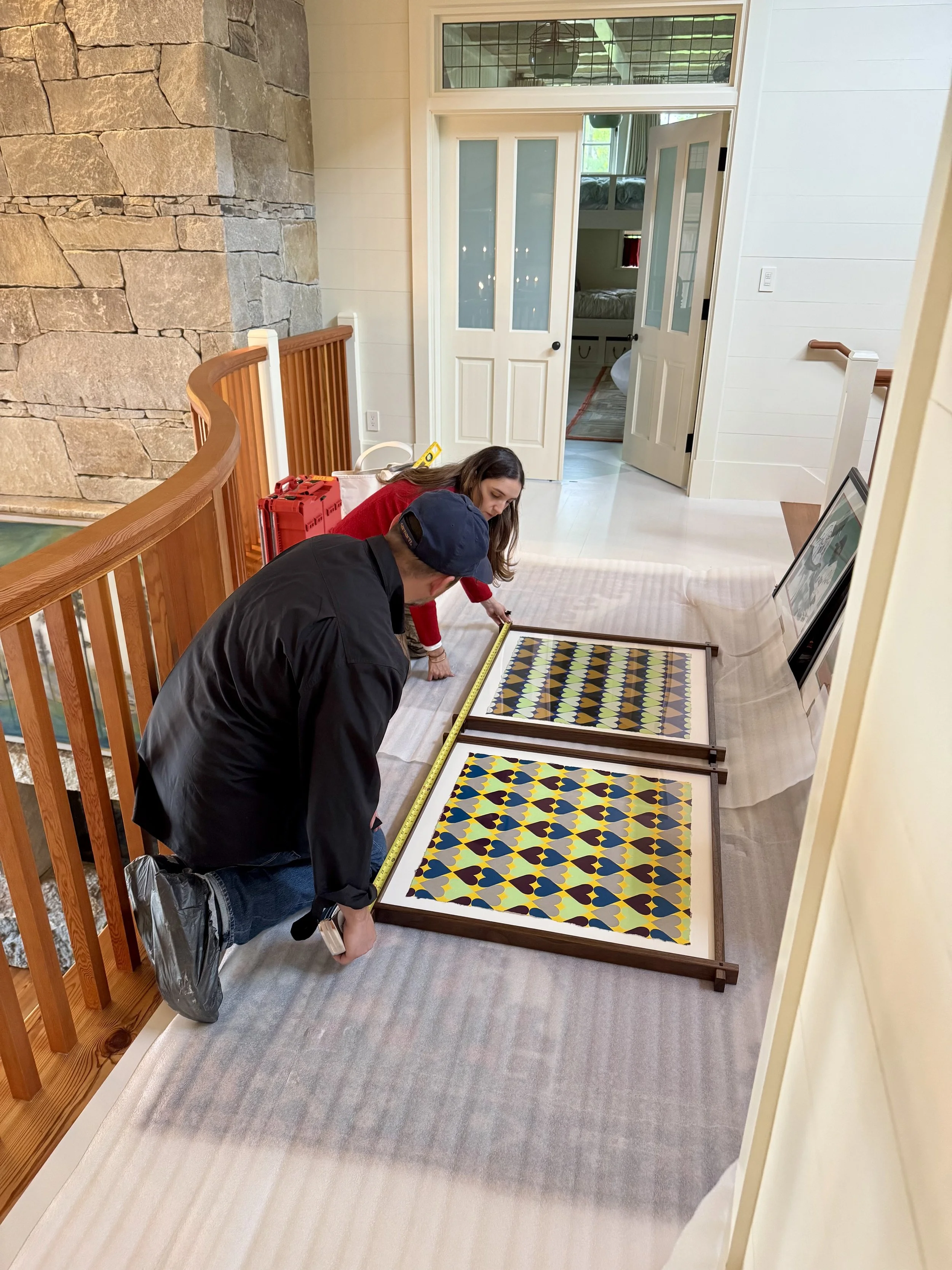 Two people measuring and working on a framed piece of art or decorative panels inside a house near a staircase and stone fireplace.