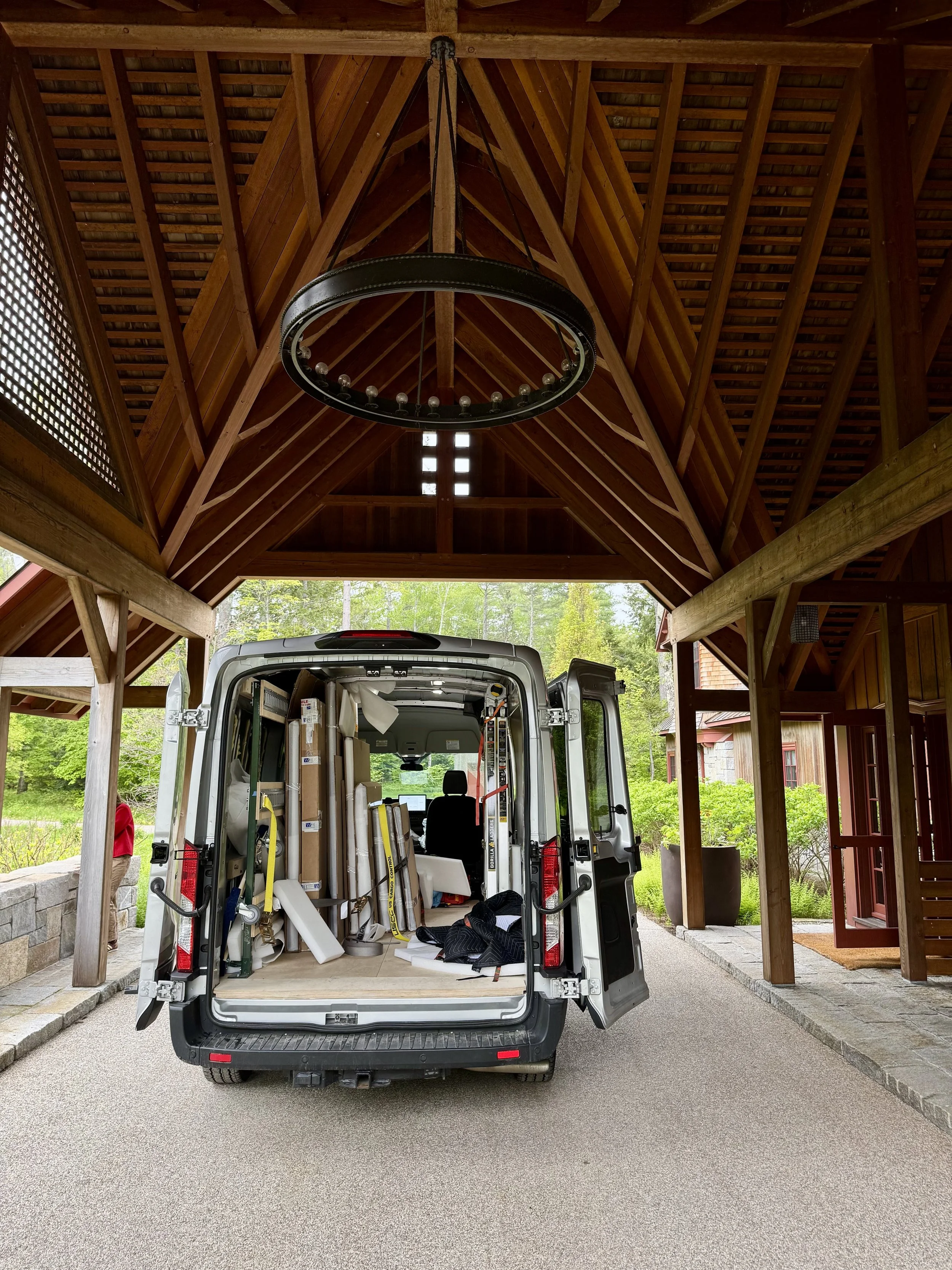 A van with its back doors open, filled with various tools and materials, parked in a covered driveway with a wooden roof and trees in the background.