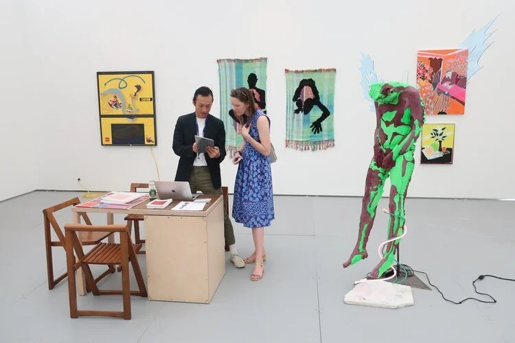Two people stand near a desk in an art gallery, looking at a tablet. The gallery displays colorful artwork on white walls, including a mixed media sculpture of a figure with wings, painted in green and purple, standing on a white platform with wires.