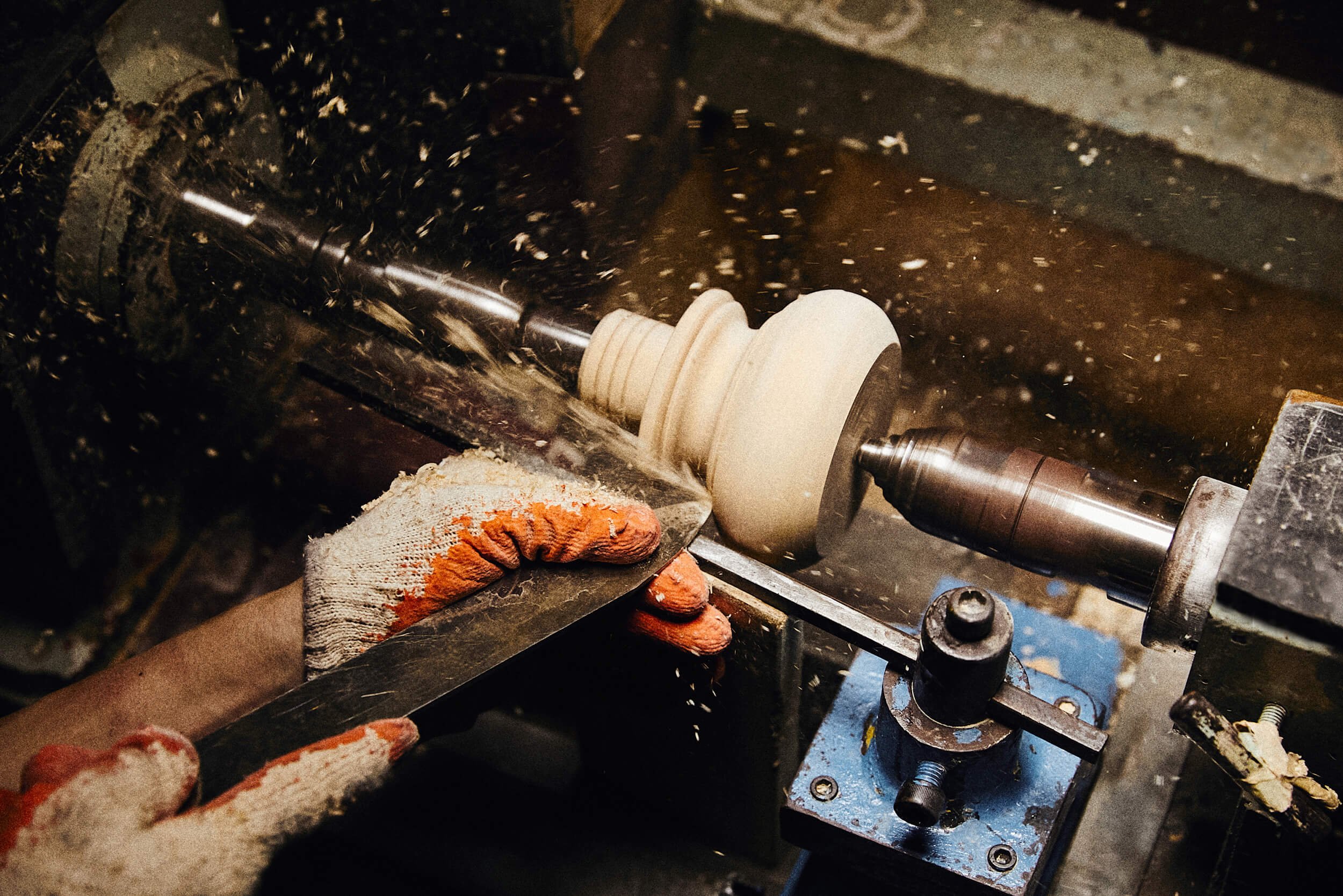 Person wearing gloves shaping a wooden object on a lathe machine, with wood shavings flying around.
