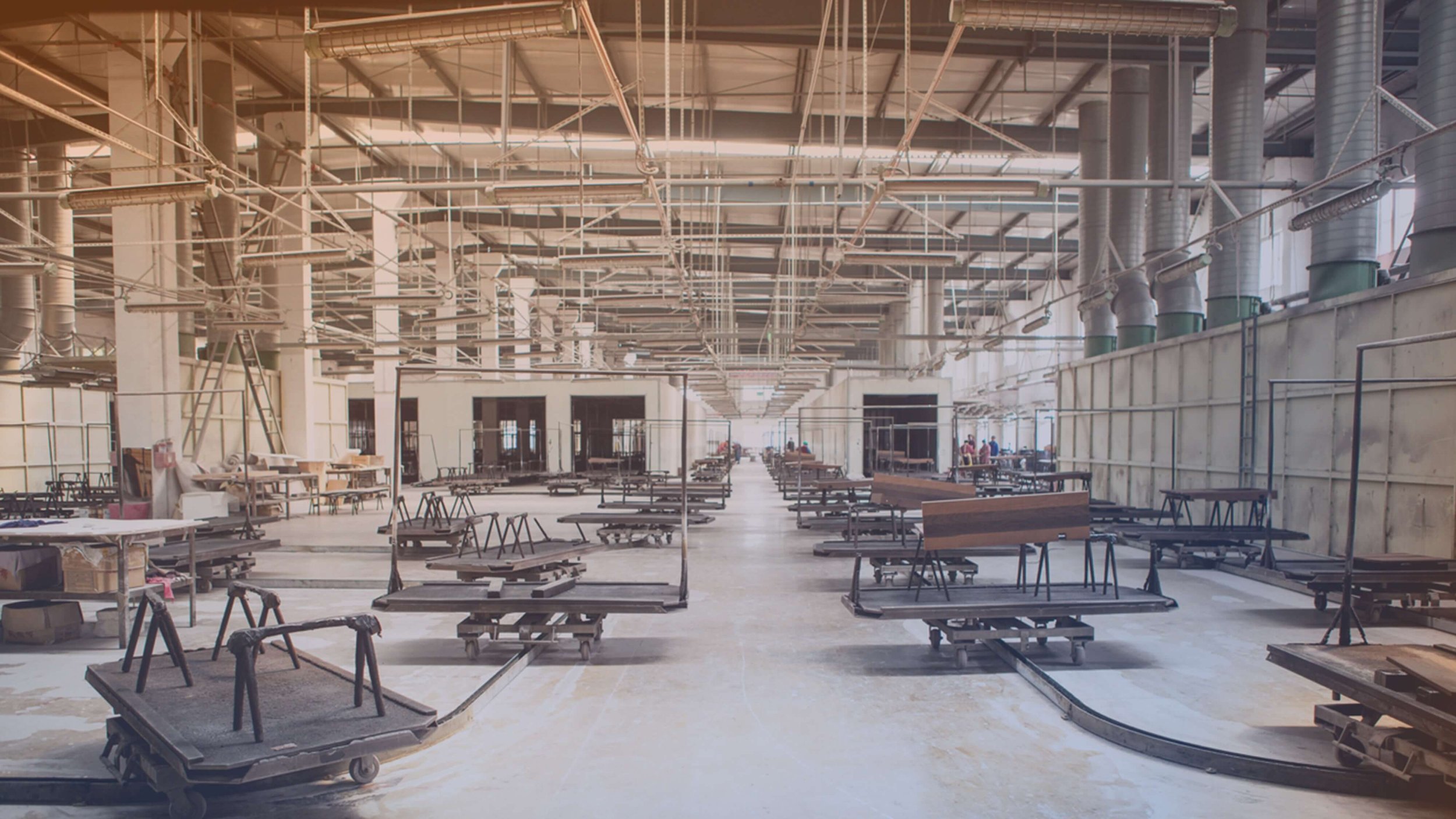 Empty industrial or manufacturing facility with rows of carts and workstations, exposed piping, and unfinished walls.