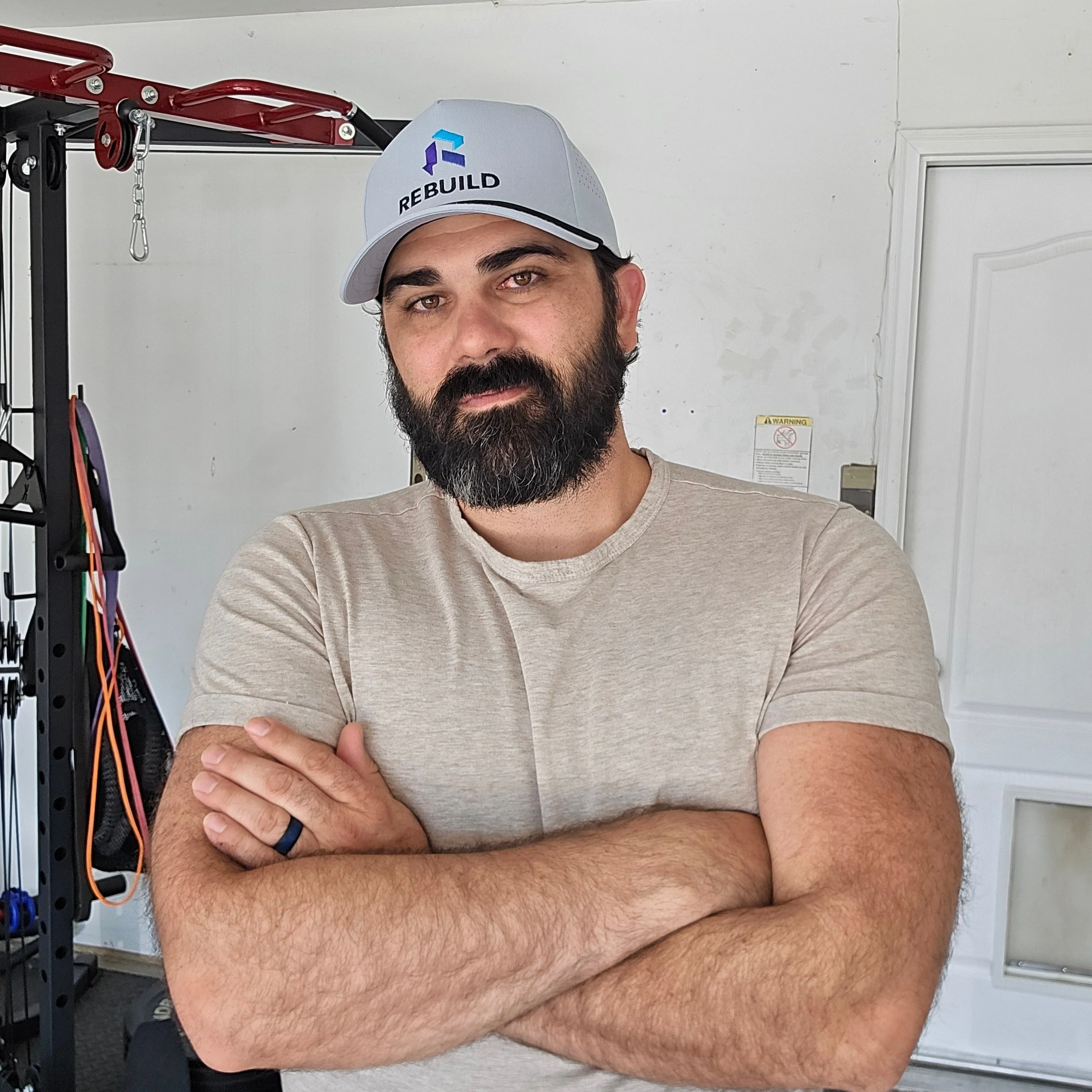 A man with a beard and short dark hair smiling, standing with arms crossed in a gym, behind him are exercise mats and fitness equipment with a sign that says "Body-Solid Built for Life."