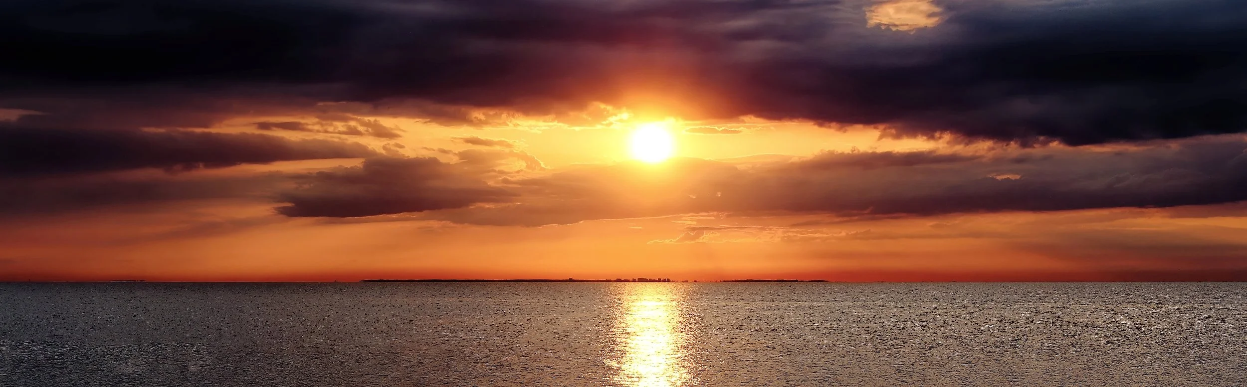 A sunset over a body of water with dark clouds in the sky, casting a reflection on the water's surface.