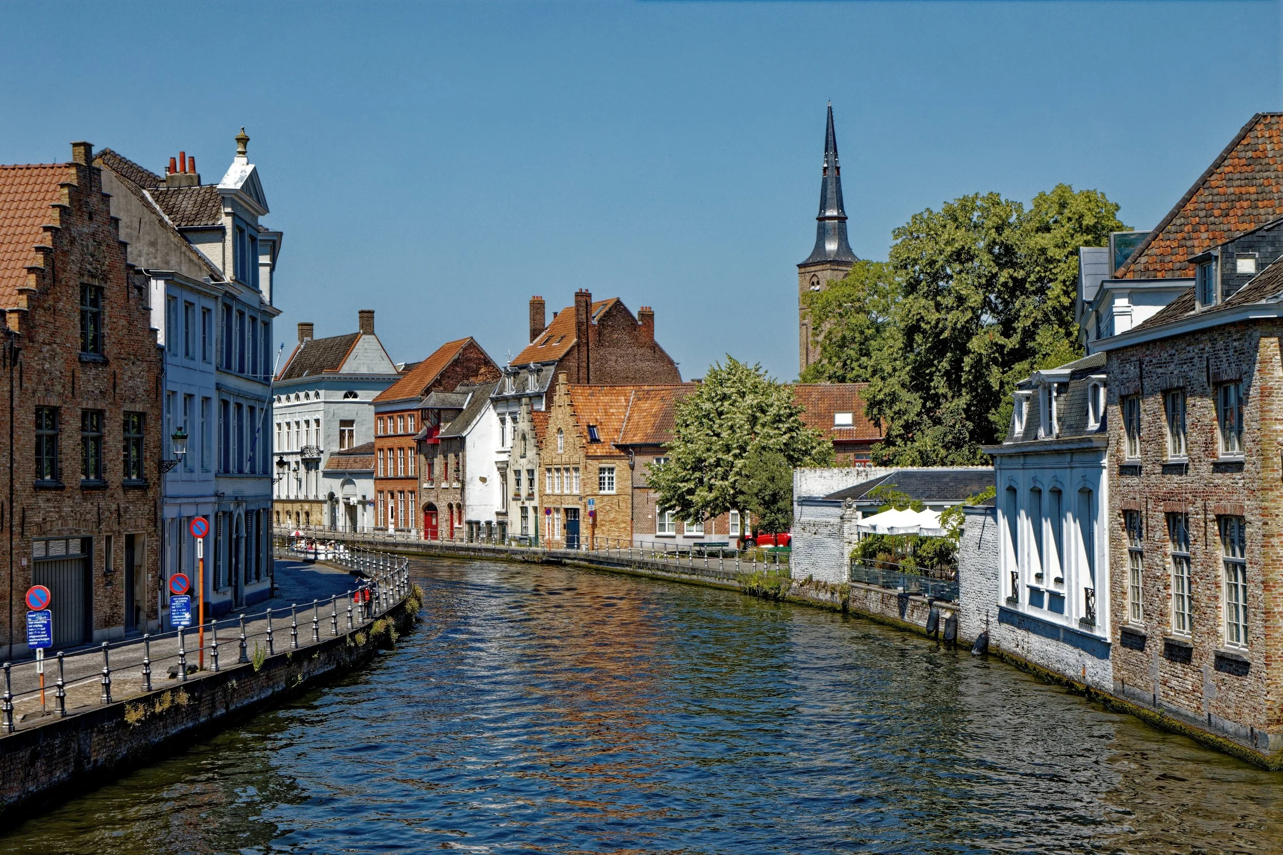 A scenic view of a canal lined with old brick and painted buildings under a clear blue sky, with some trees and a church steeple in the background.
