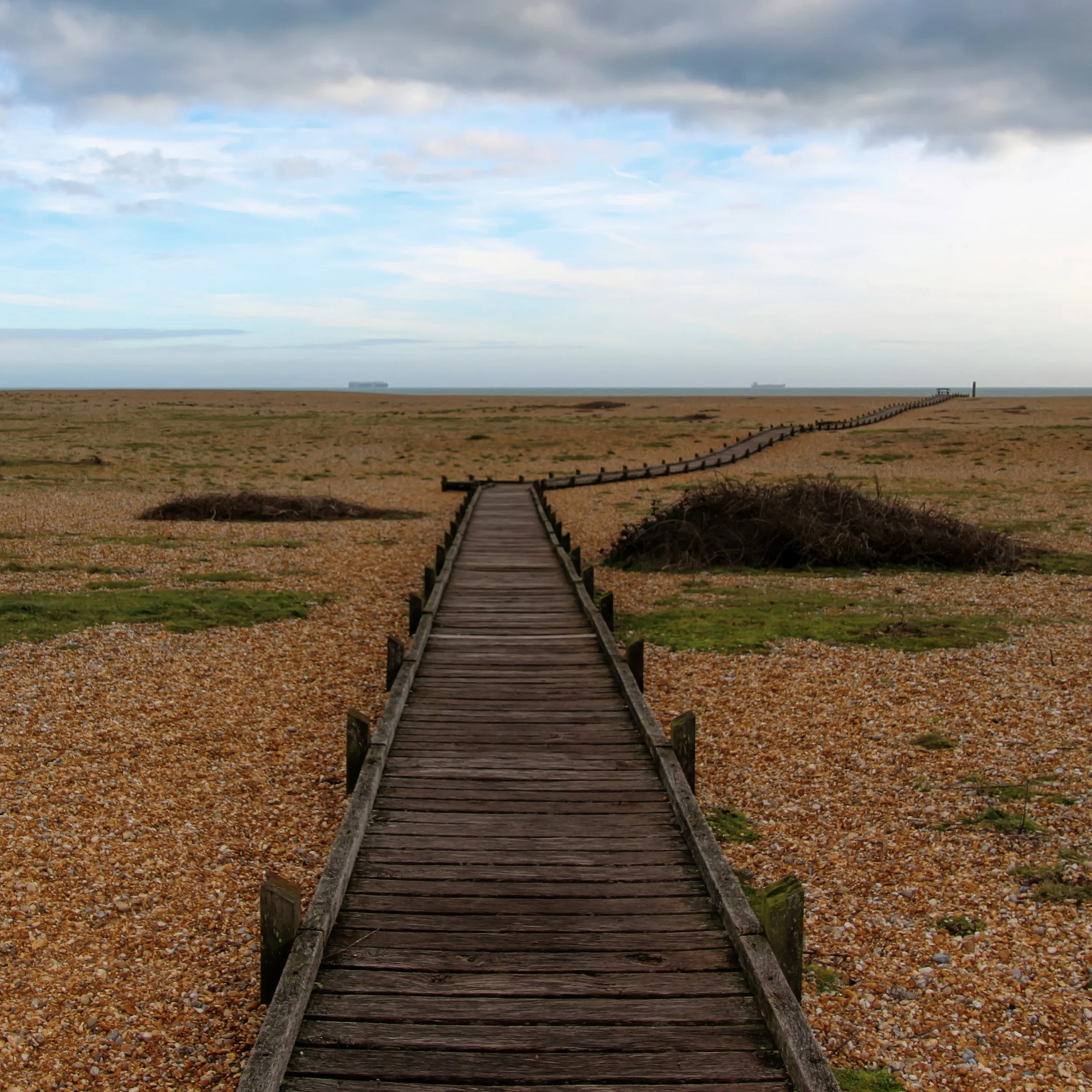 A wooden boardwalk extends over a pebbled beach towards the sea, with a cloudy sky overhead and ships visible on the horizon.