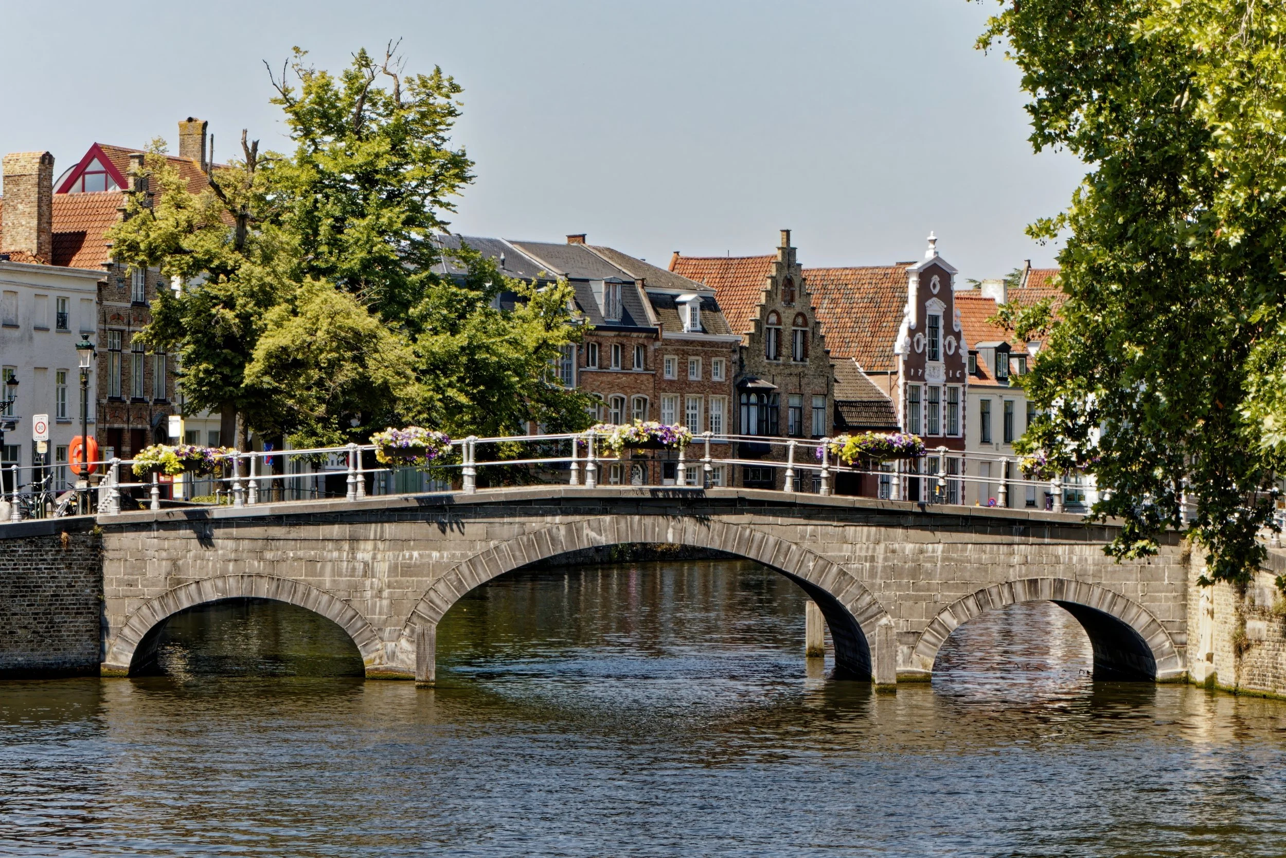 A stone bridge with arches over a river, with historic European-style buildings and trees in the background, and flower boxes on the bridge railing.