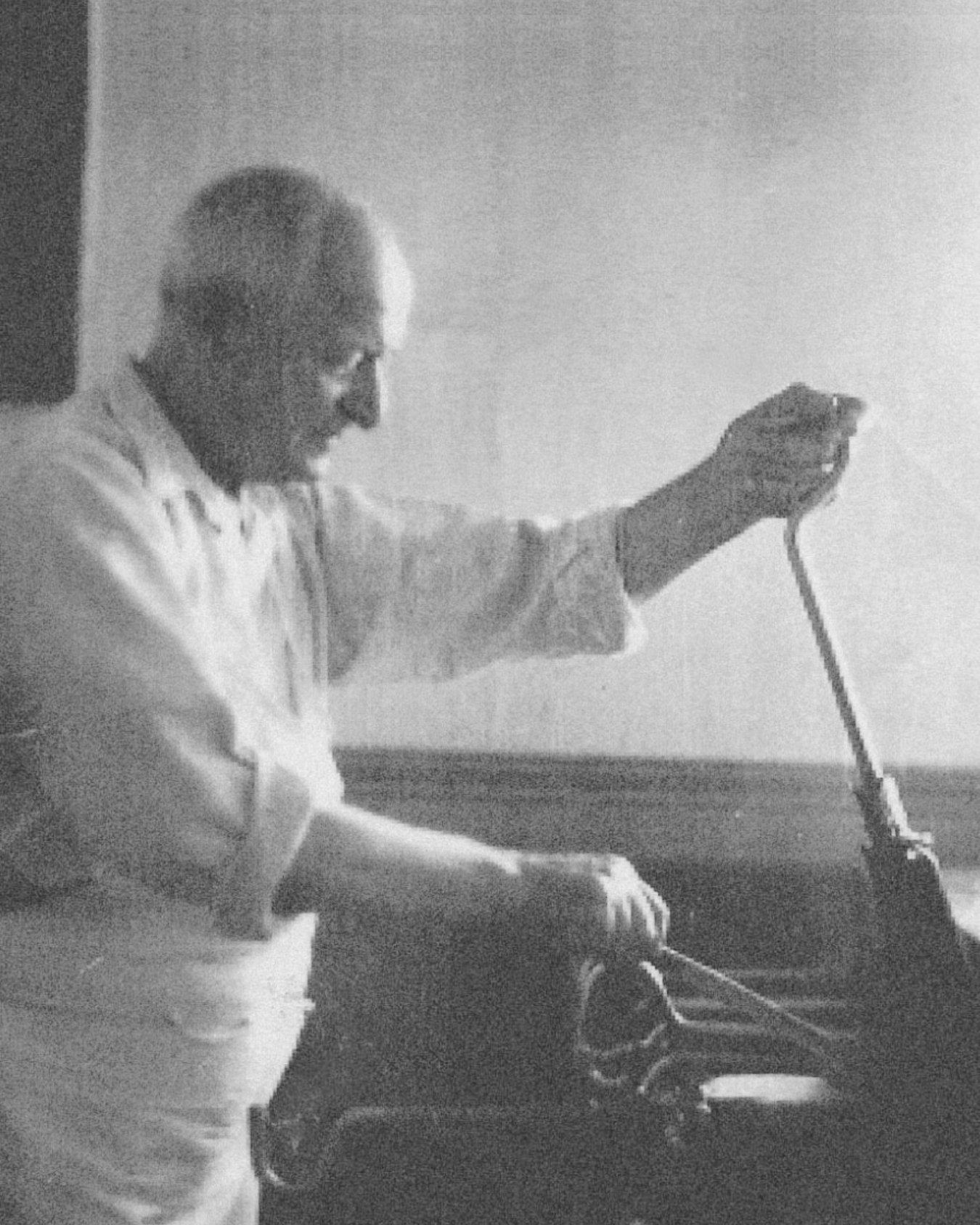 Un homme âgé souriant en tenue de chef, avec une moustache, utilisant un microphone ou un autre appareil de communication dans un environnement intérieur.