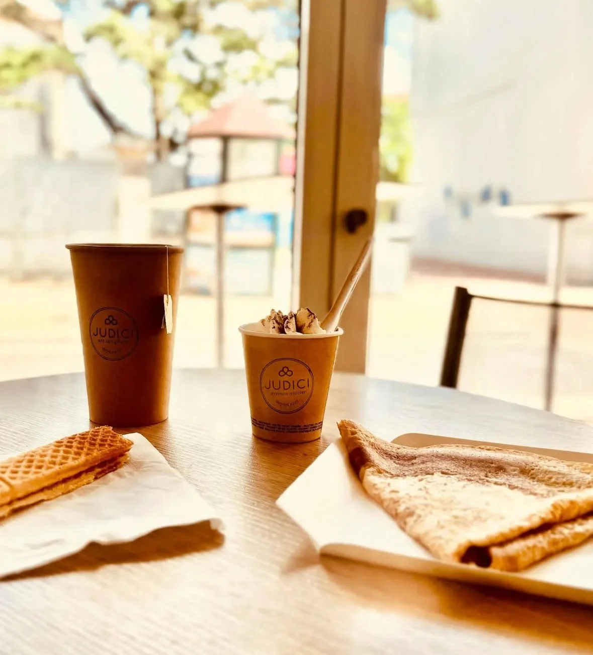 Une table avec deux boissons chaudes, une gaufrette artisanale et une crêpe, dans un intérieur avec une grande fenêtre laissant entrer la lumière naturelle.