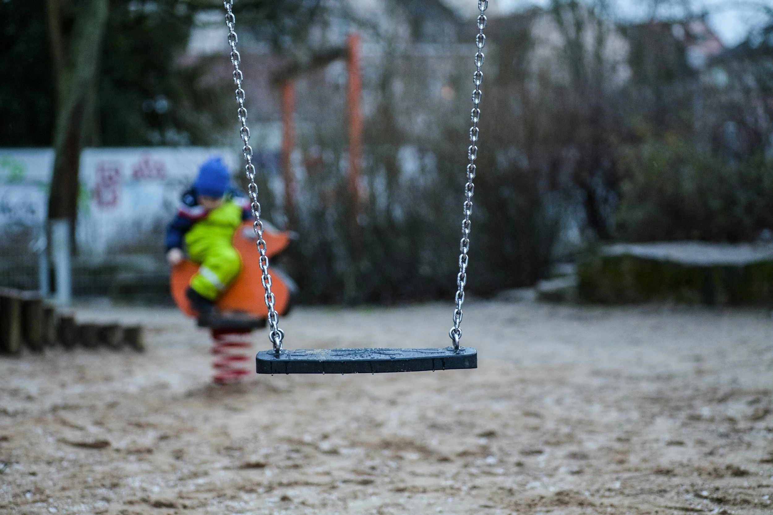 Empty swing seat in a playground with a blurred child on a spring rider in the background.