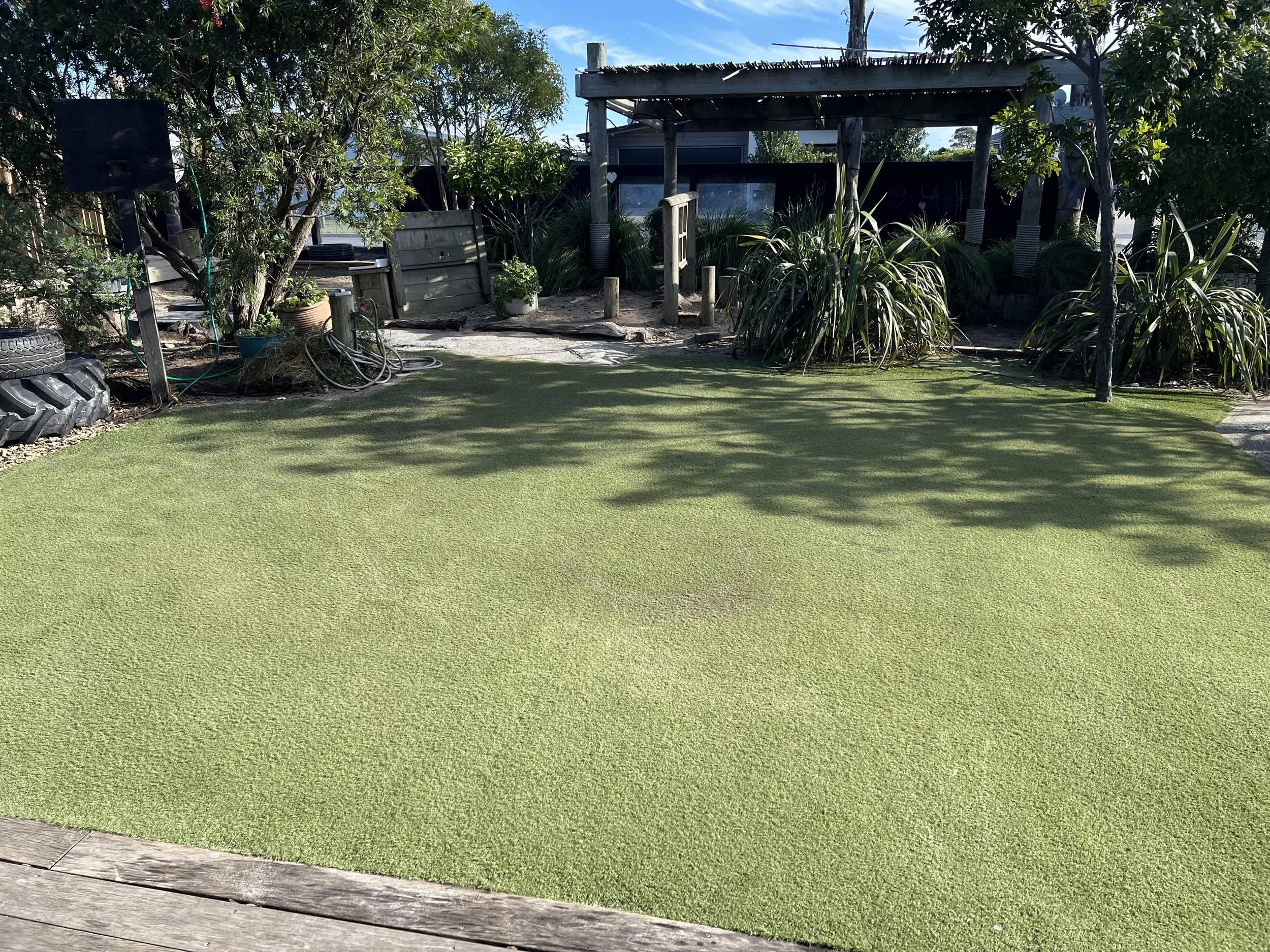 Backyard with a patch of artificial grass, trees, shrubs, a small wooden pergola, and outdoor furniture.