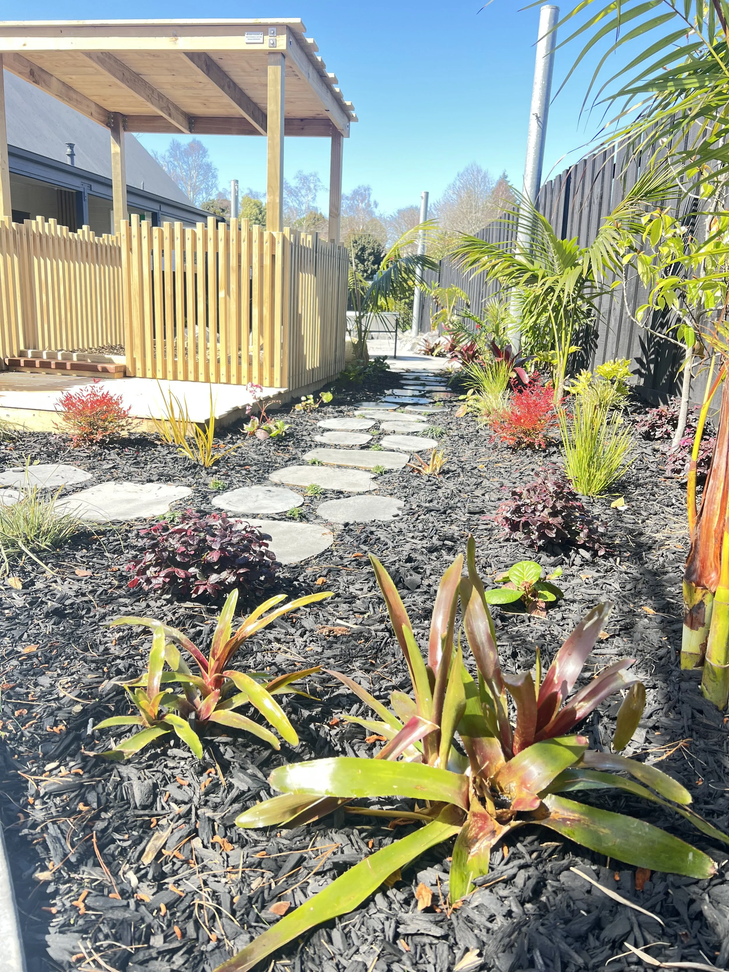 A landscaped backyard garden with a stone pathway, tropical plants, mulch, a wooden fence, and a deck area under construction.