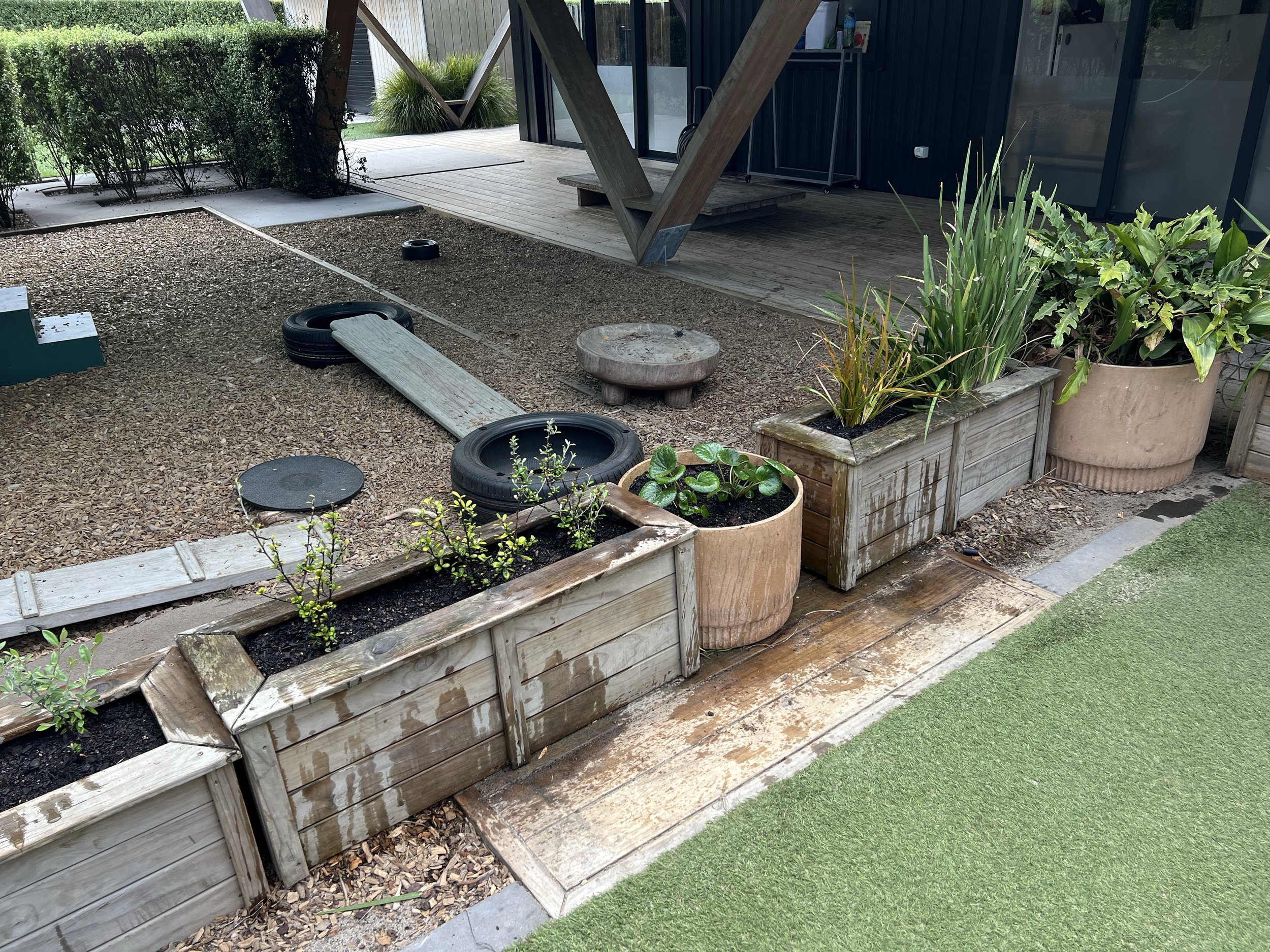 Garden at backyard with wooden planters, tires, and stepping stones, adjacent to a wooden deck and artificial grass.