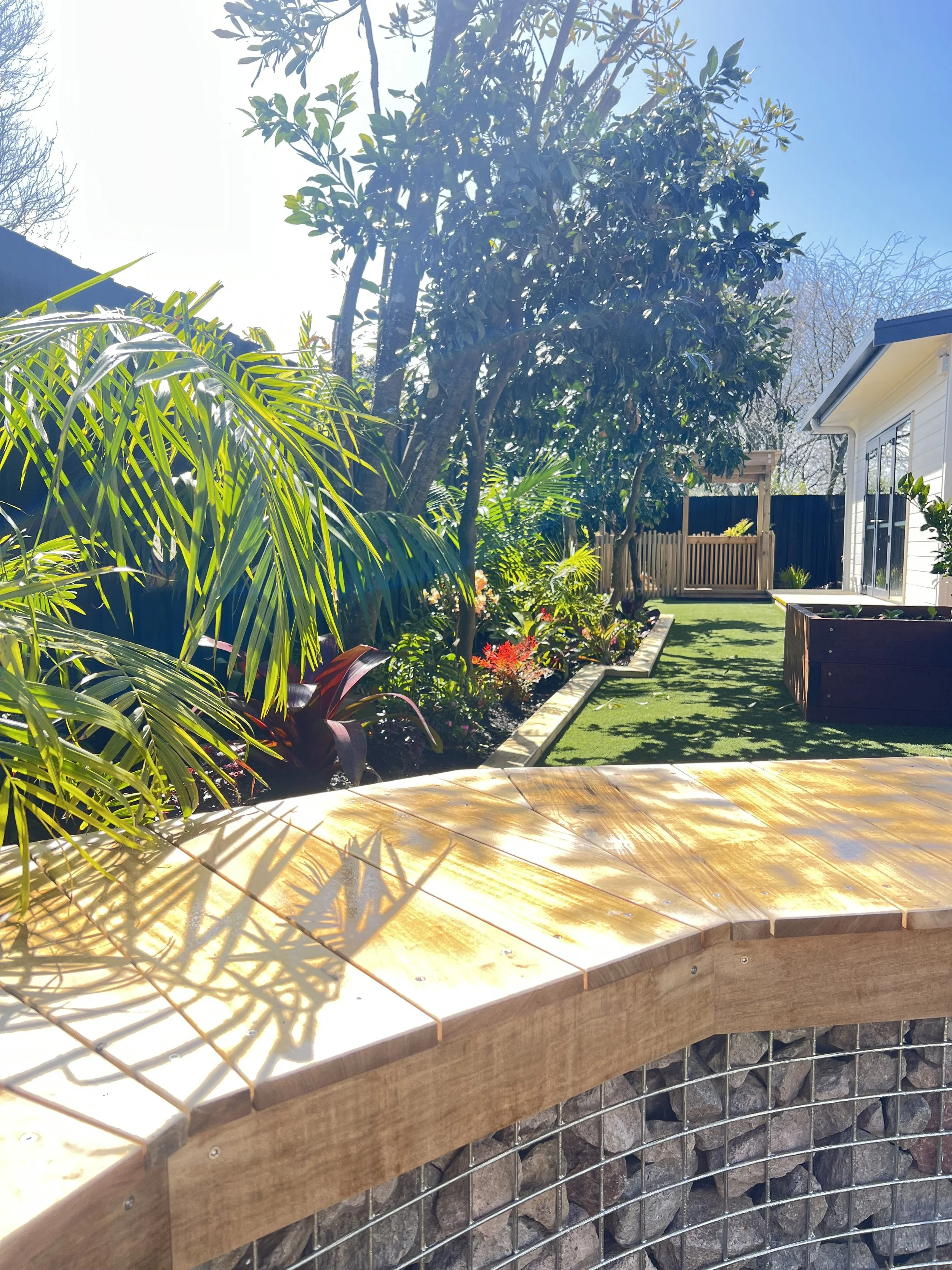 Backyard garden with lush green trees and plants, a small grassy area, and a white house with sliding glass doors, all under a sunny blue sky.