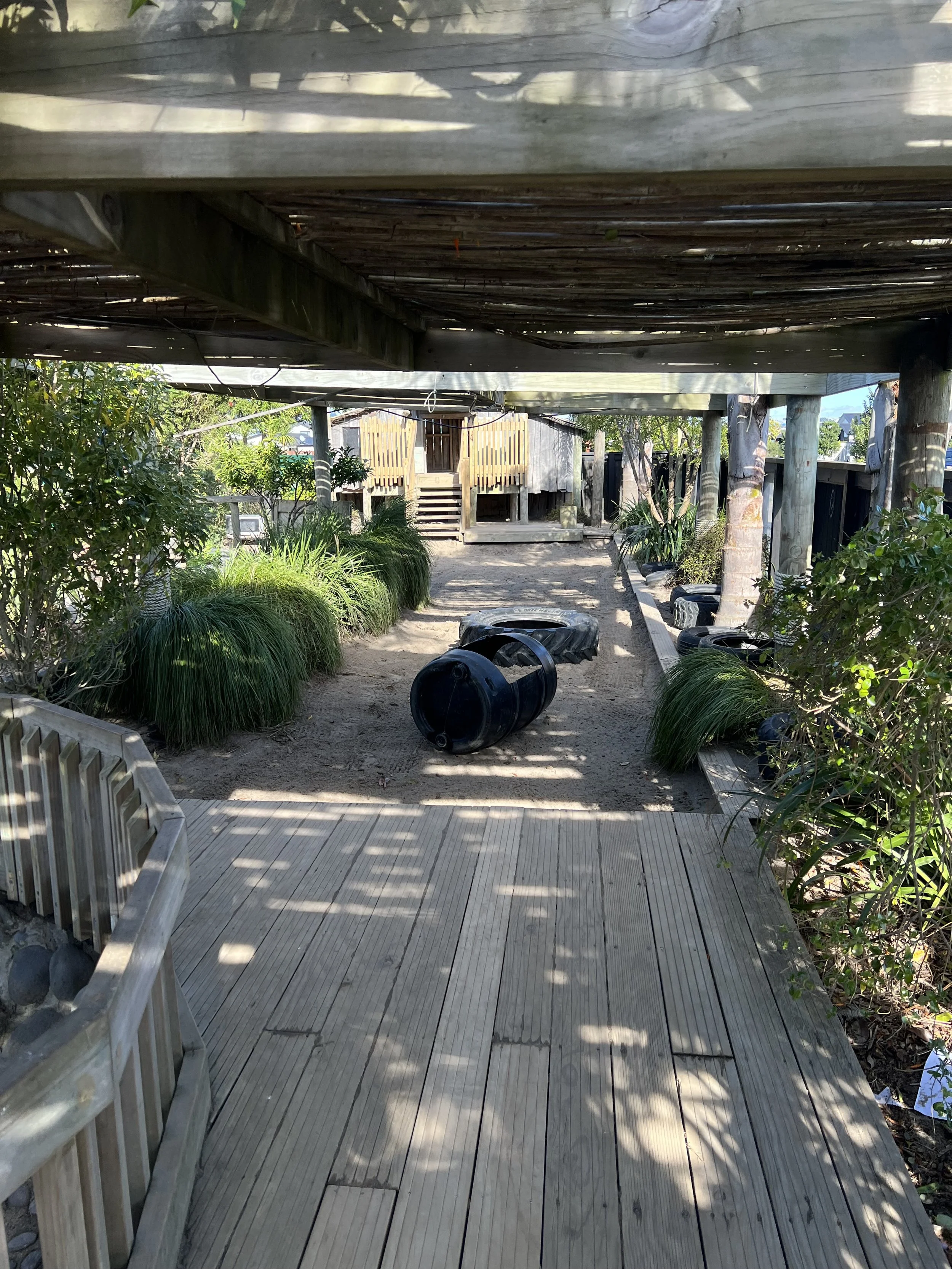 A raised wooden deck leads to a sandy play area with black tires and plastic tubing, surrounded by green plants and trees, with a small wooden structure in the background.