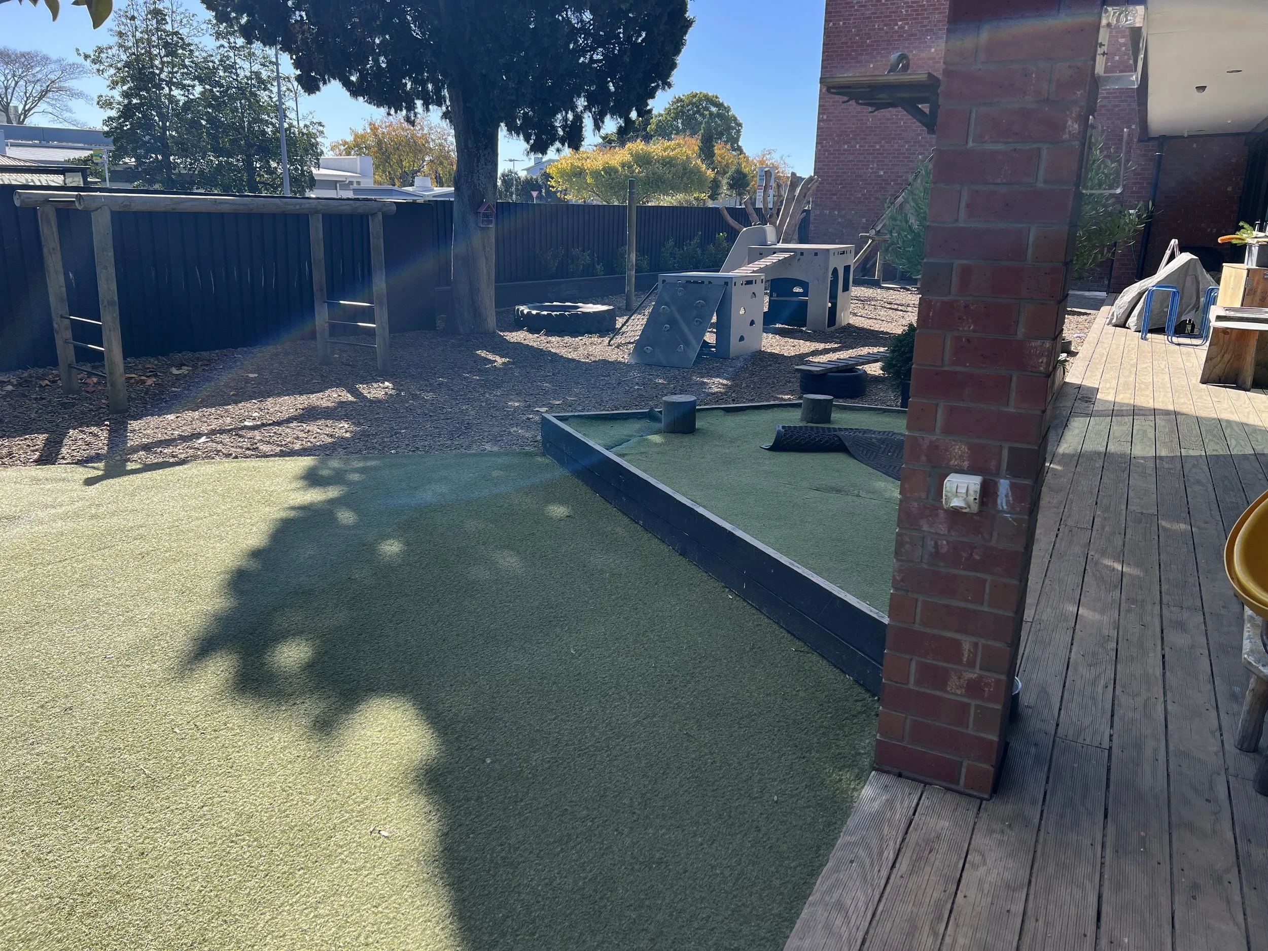 Backyard with artificial grass, wooden deck, climbing wall, and play structures under sunny sky.