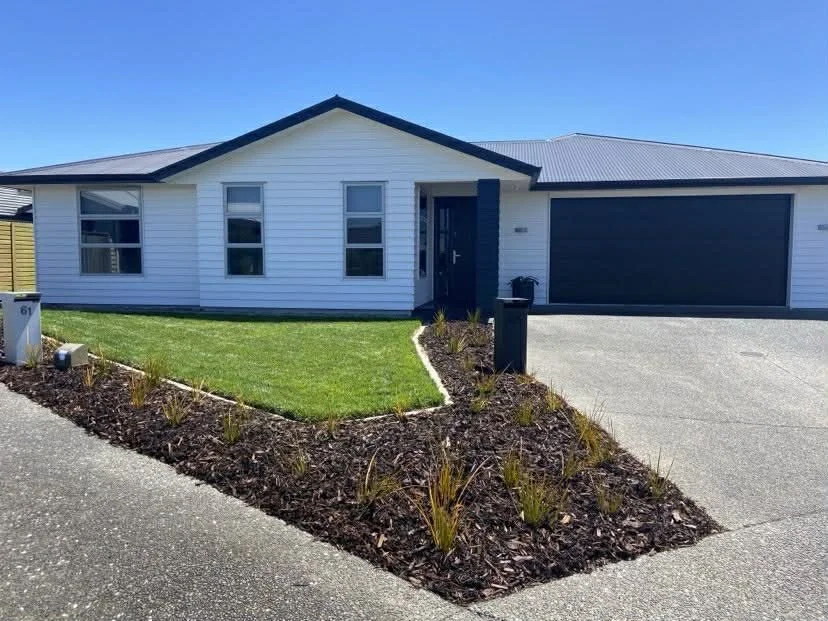 Modern house with white siding, a dark gray garage door, a small front yard with green grass, and a flower bed with mulch and plants bordering the driveway.