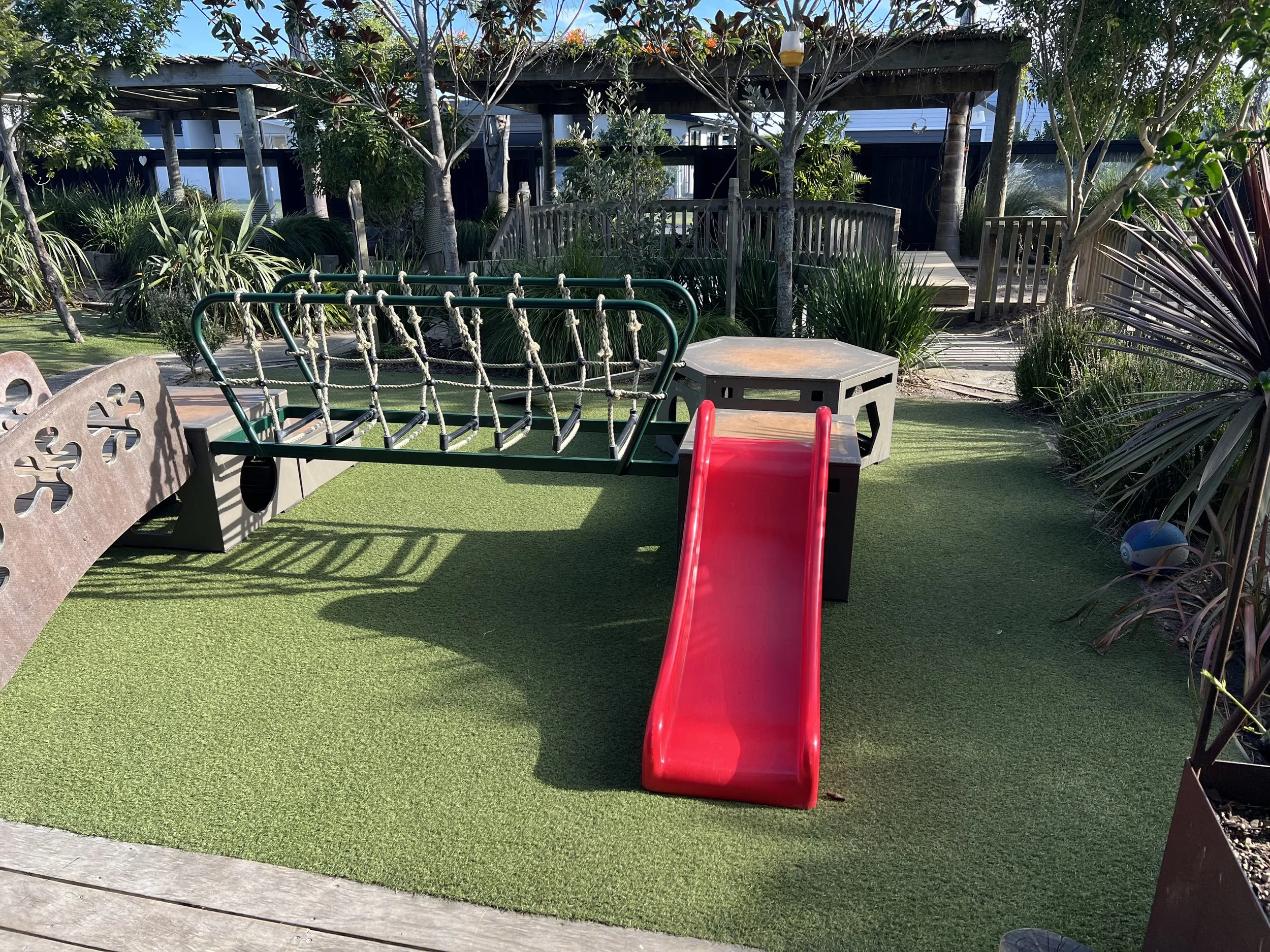 Children's outdoor playground with red slide, climbing net, and sandbox area, surrounded by green artificial turf and plants.