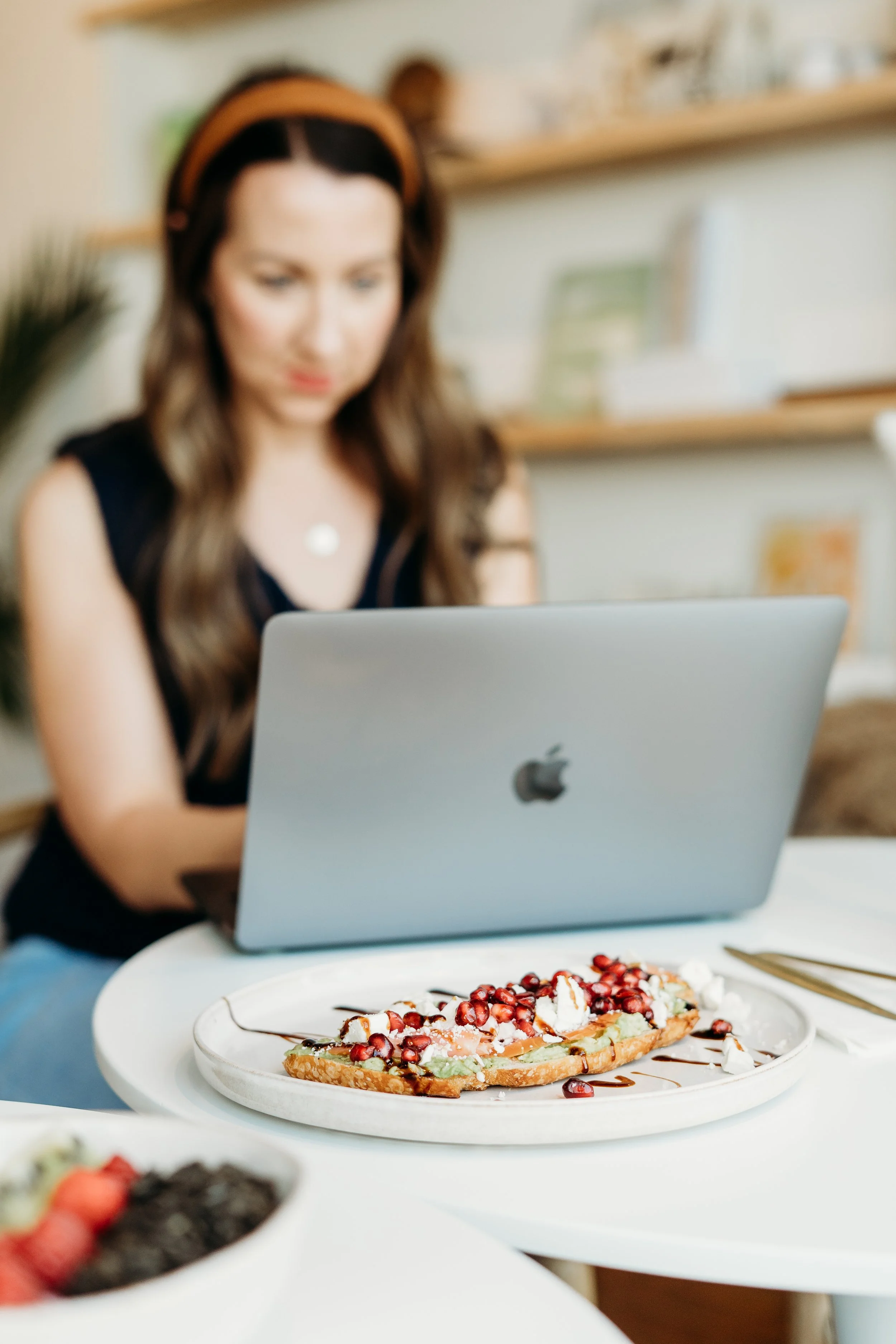 A woman sitting at a table with a silver MacBook laptop in front of her, and a pastry topped with pomegranate seeds, whipped cream, and chocolate drizzle on a plate.