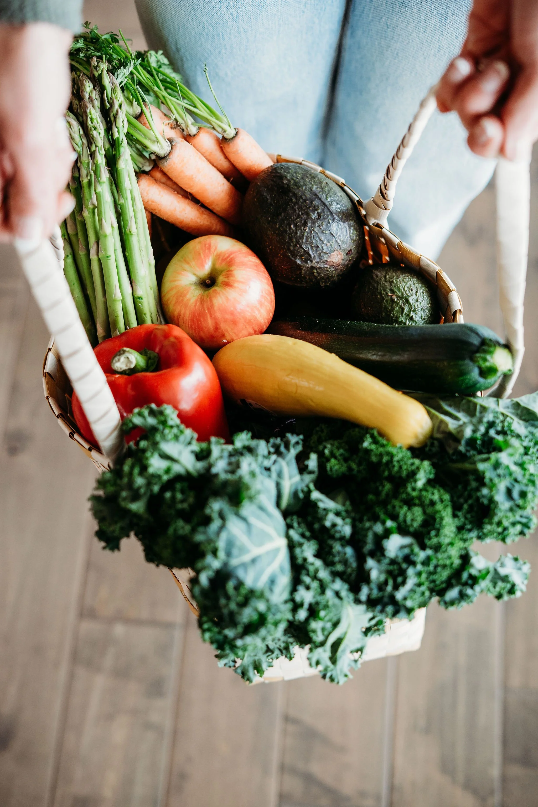 A basket of fresh vegetables including asparagus, carrots, apples, avocados, zucchini, yellow squash, red bell pepper, kale, and broccoli.